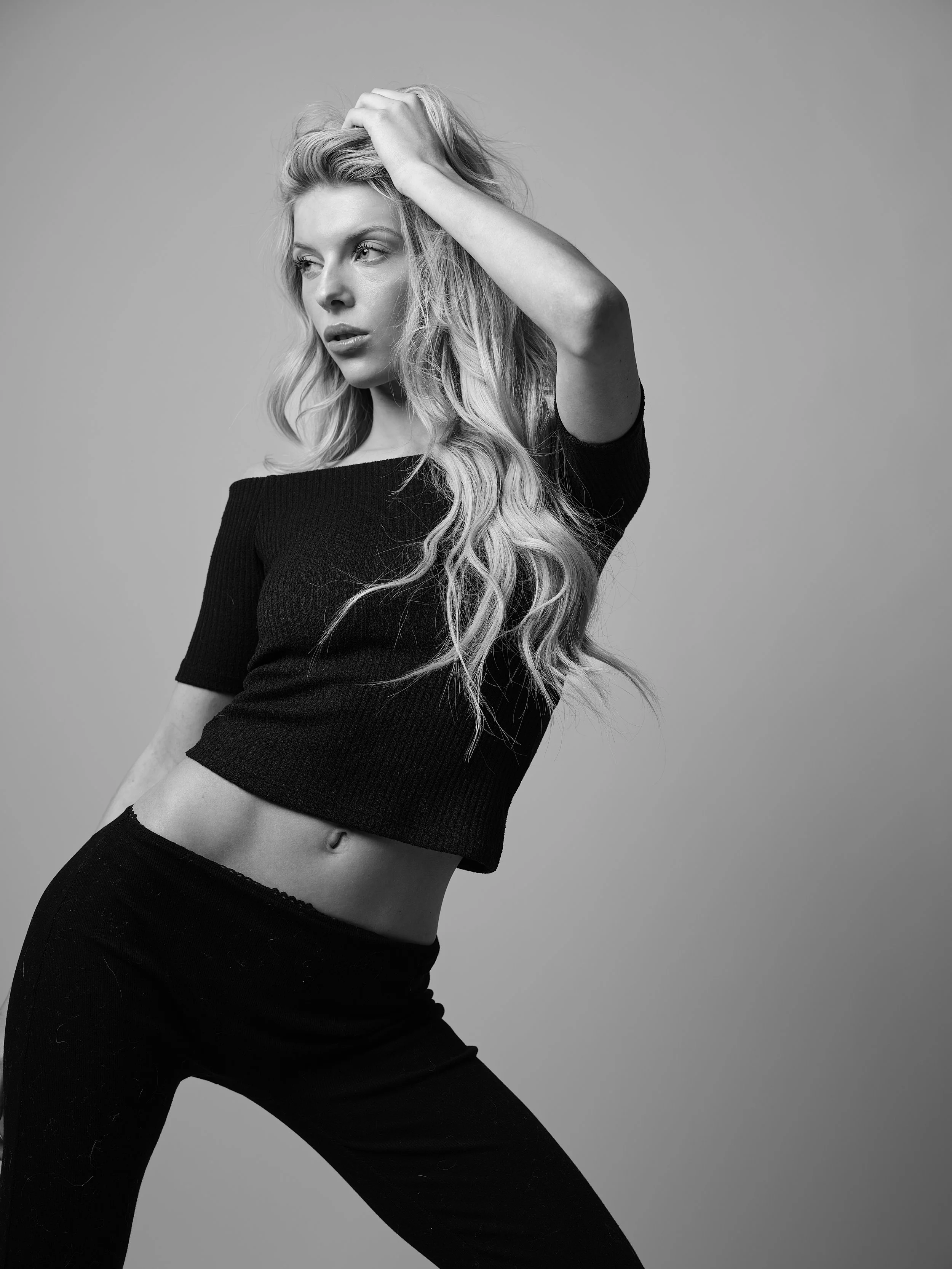 Black-and-white photo of a young woman with long wavy hair wearing a black crop top and black pants. She is posing with one hand on her head and looking to the side, against a plain background.