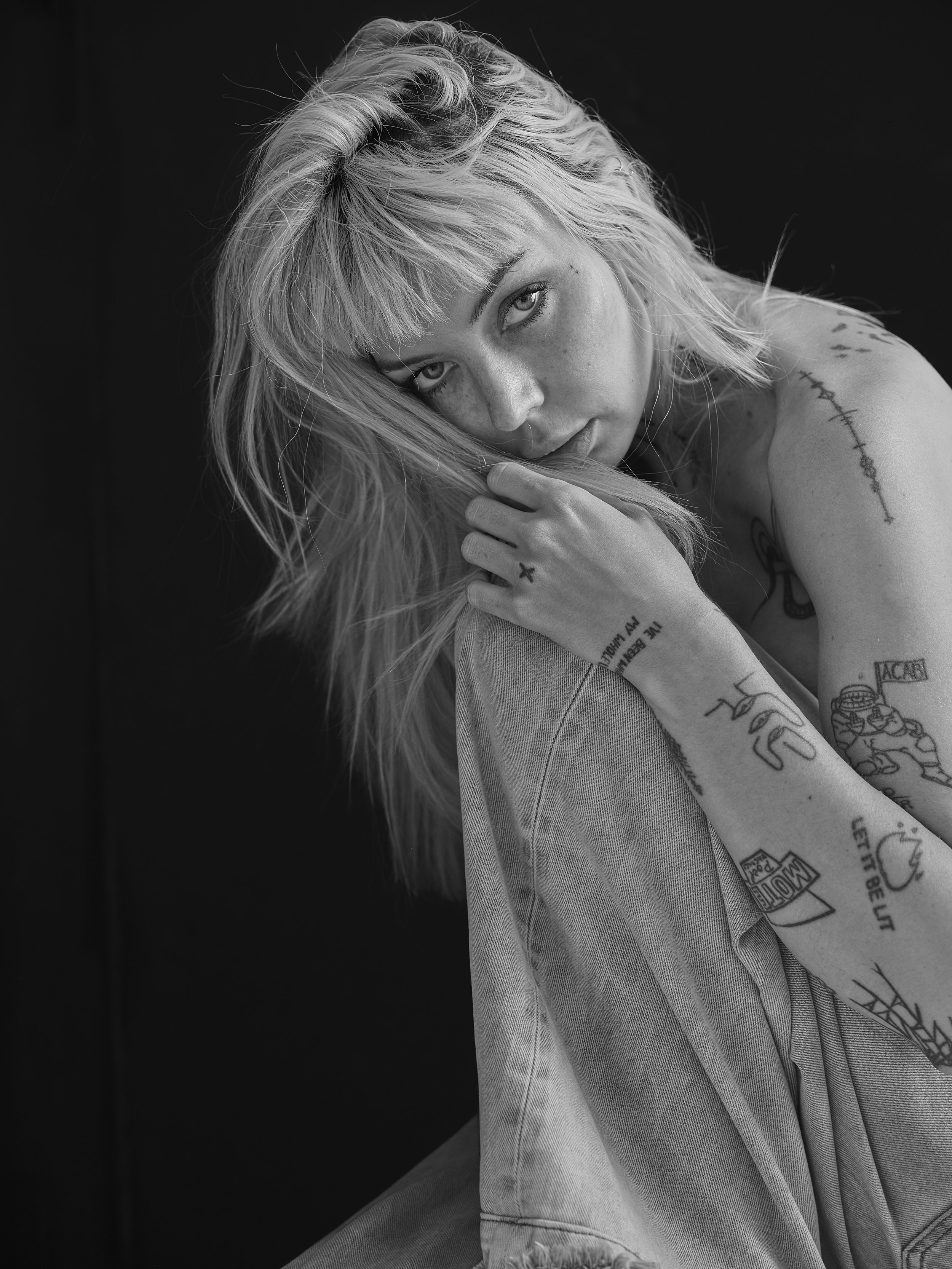 A black-and-white portrait of a young woman with blonde hair and tattoos on her arms, resting her head on her hand against a dark background.