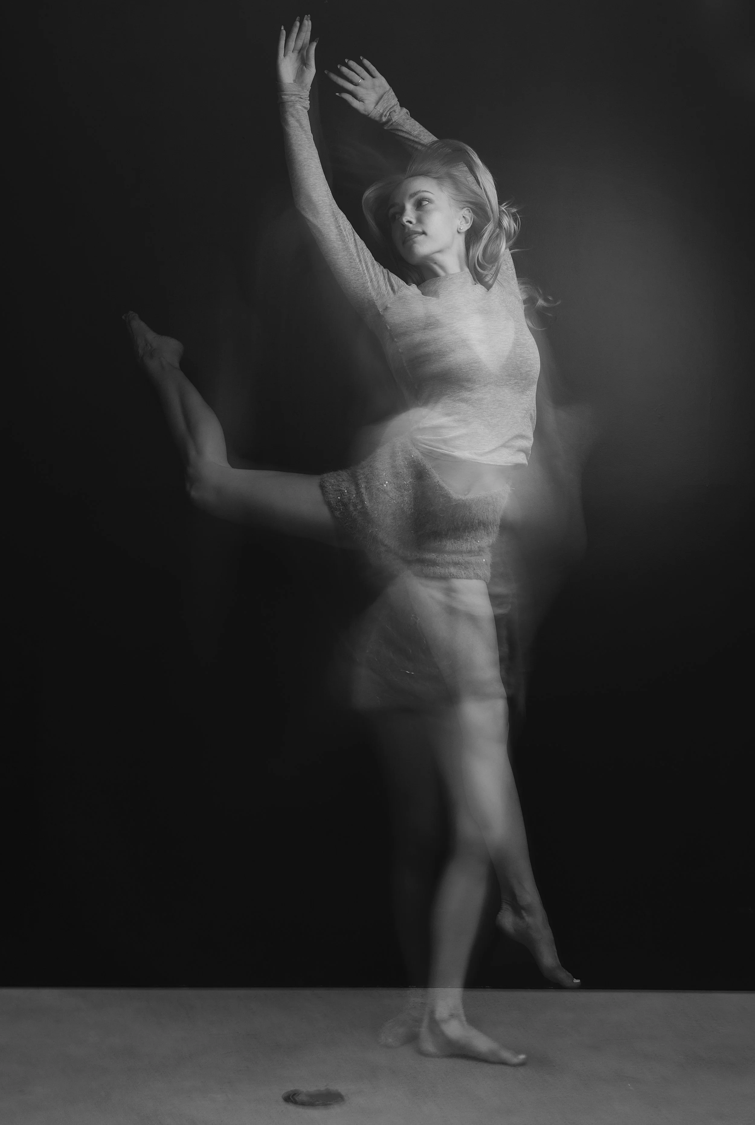 Black and white photo of a young woman performing a ballet dance on stage, her arms raised above her head, one leg bent and her toes pointed on the floor.