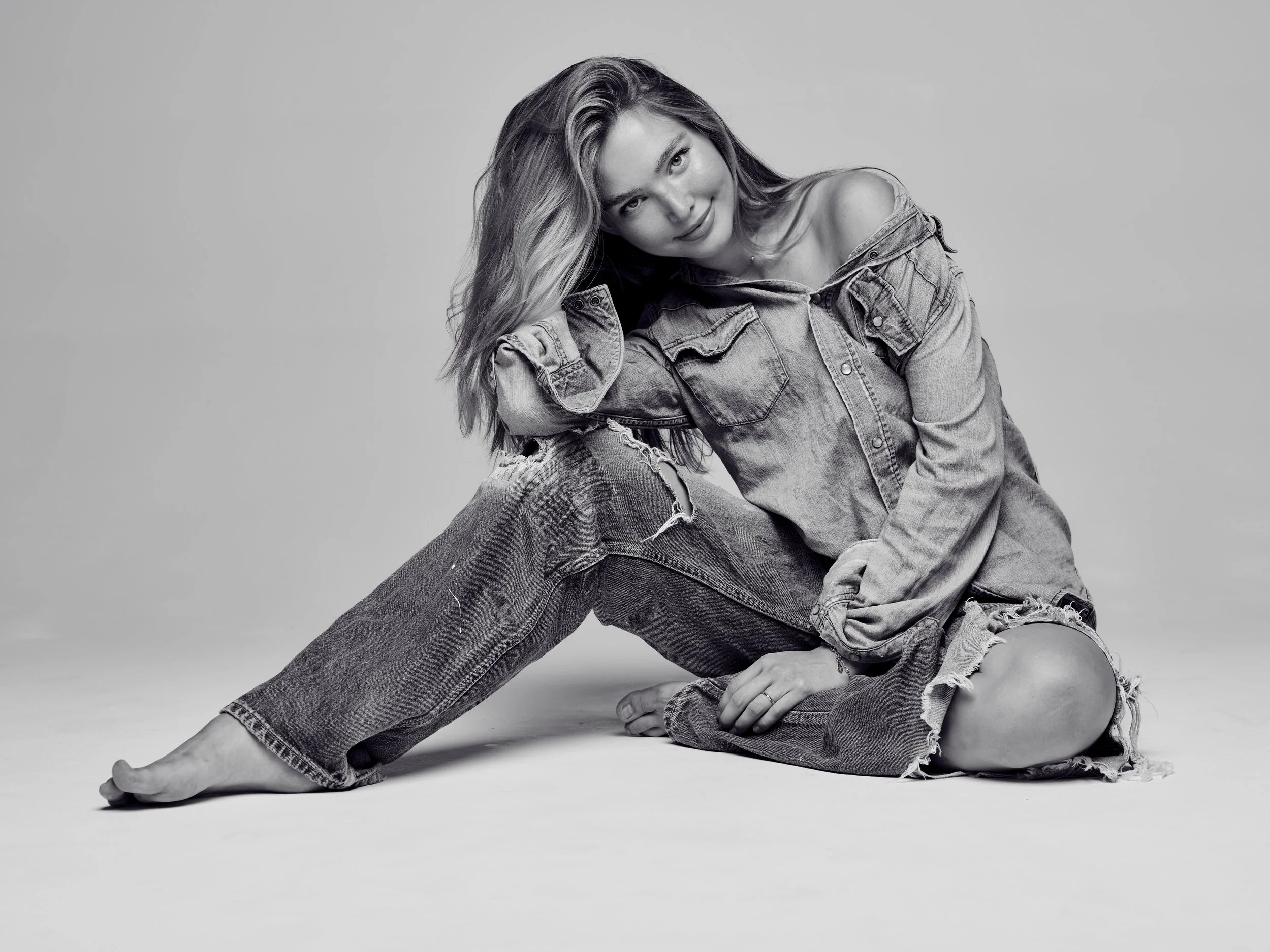 Black and white photo of a woman with long wavy hair, smiling, wearing a denim jacket over a shirt, sitting barefoot on the floor with one leg extended and the other bent, leaning on one arm against her knee.