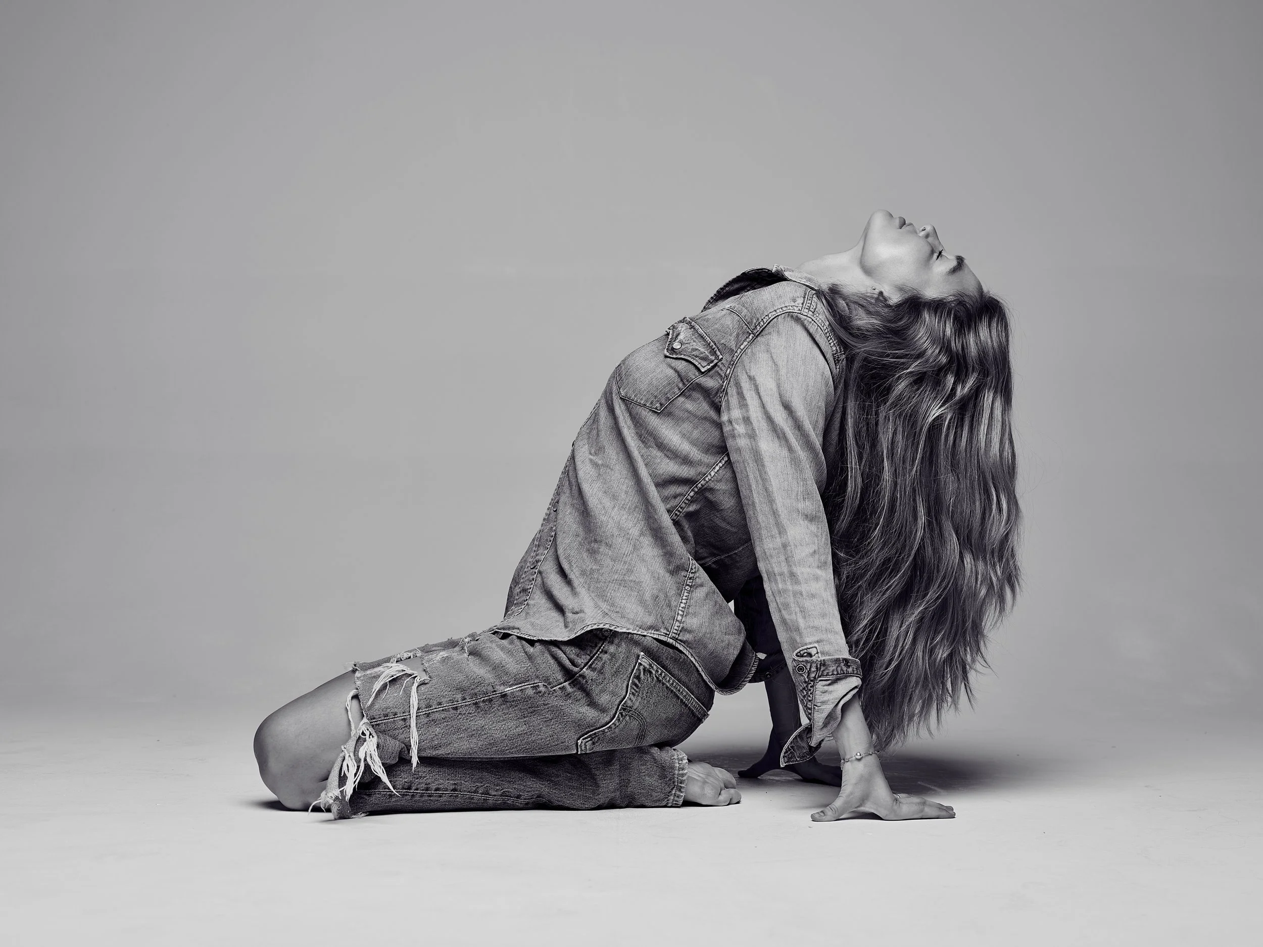 A young woman with long hair, wearing a denim jacket and ripped jeans, poses on the ground with her back arched and head tilted back, against a plain gray background.