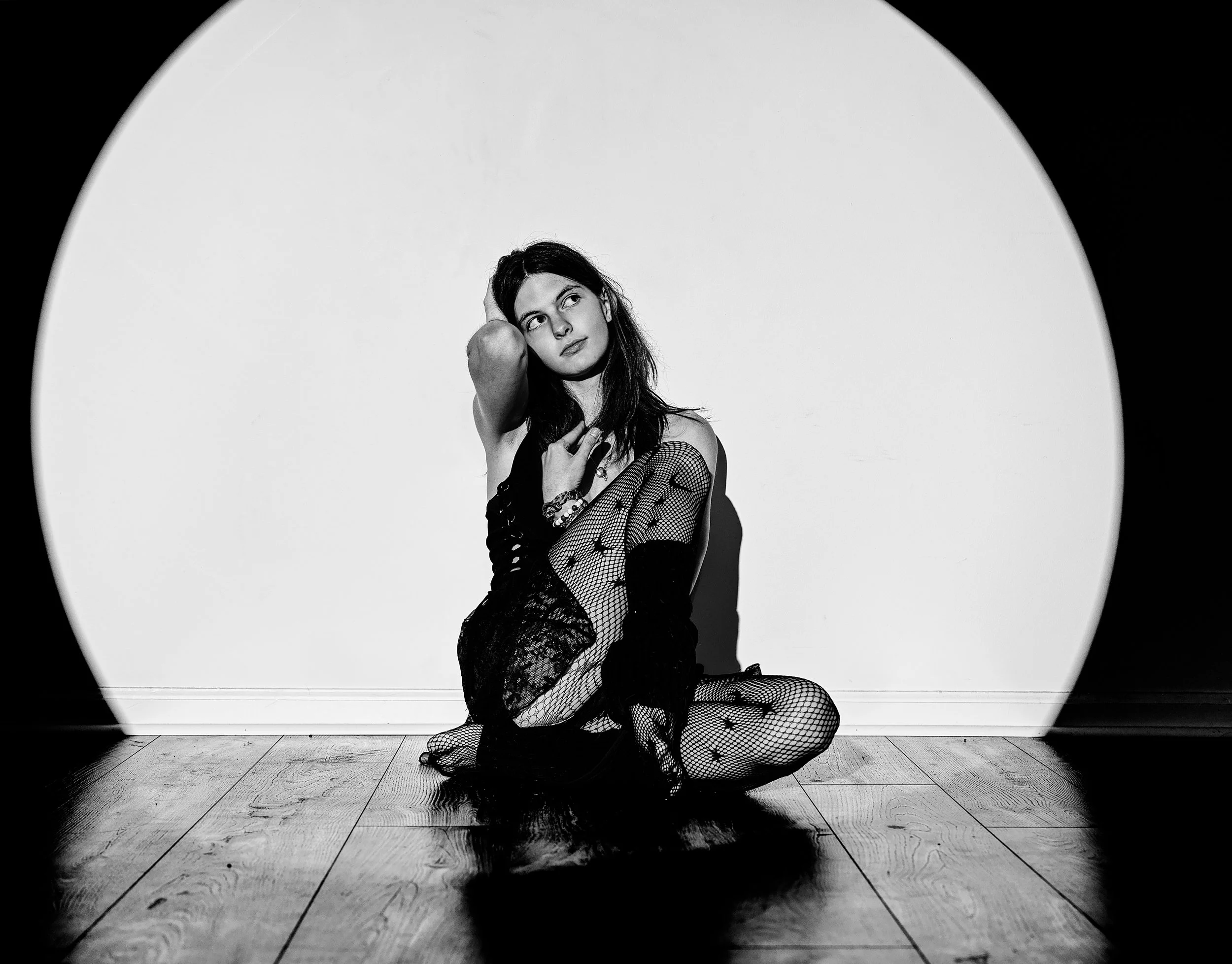 A woman with long dark hair sitting on a hardwood floor surrounded by a large circular light or spotlight, wearing fishnet stockings and a black outfit, with her hand on her head and her gaze directed upward.