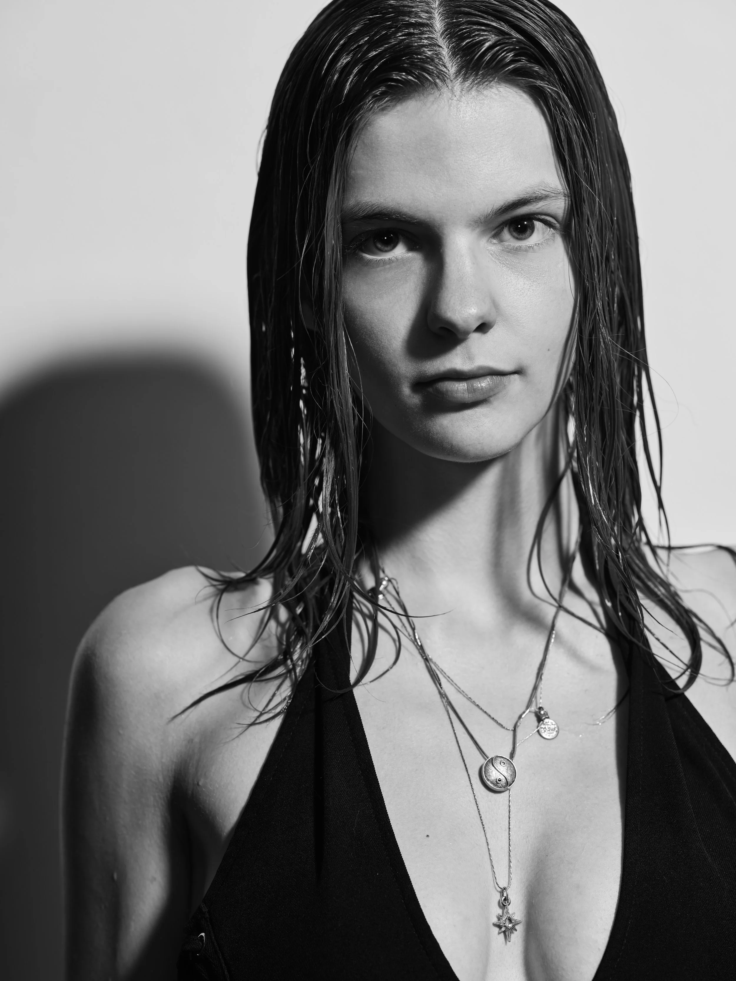 Black and white portrait of a woman with wet hair, wearing a sleeveless top and layered necklaces with crescent and star pendants, looking directly at the camera.