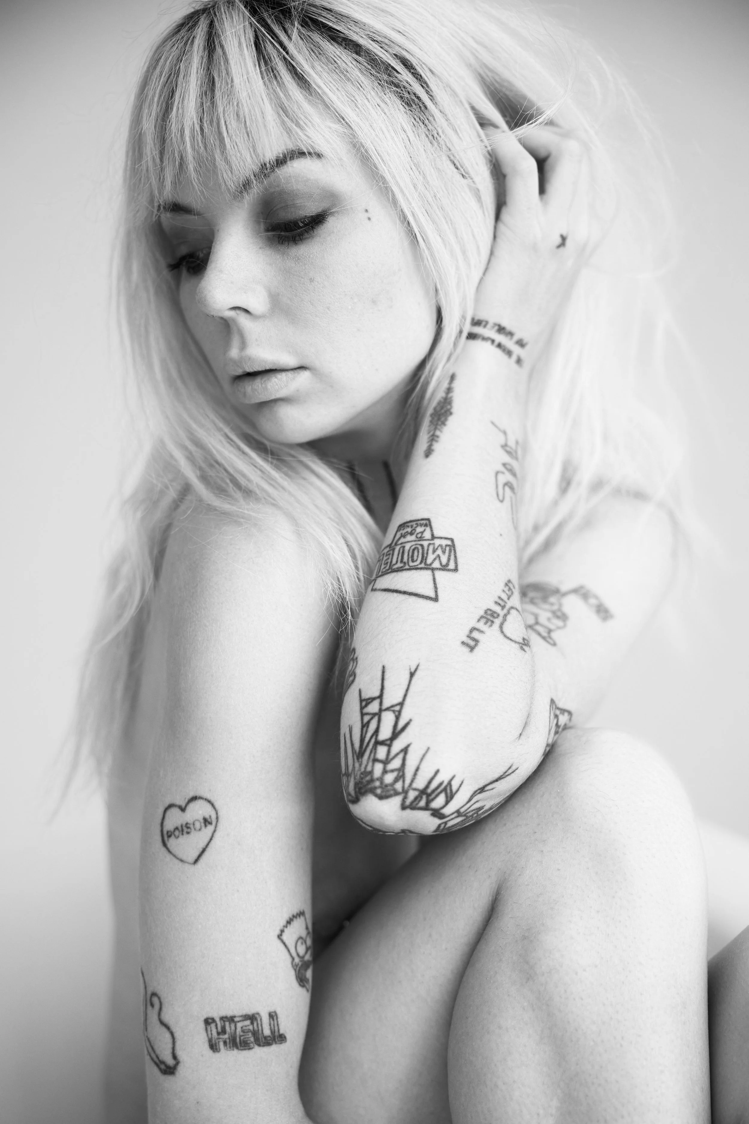 A black and white photo of a woman with long blonde hair and tattoos on her arm and shoulder, touching her hair and looking down.