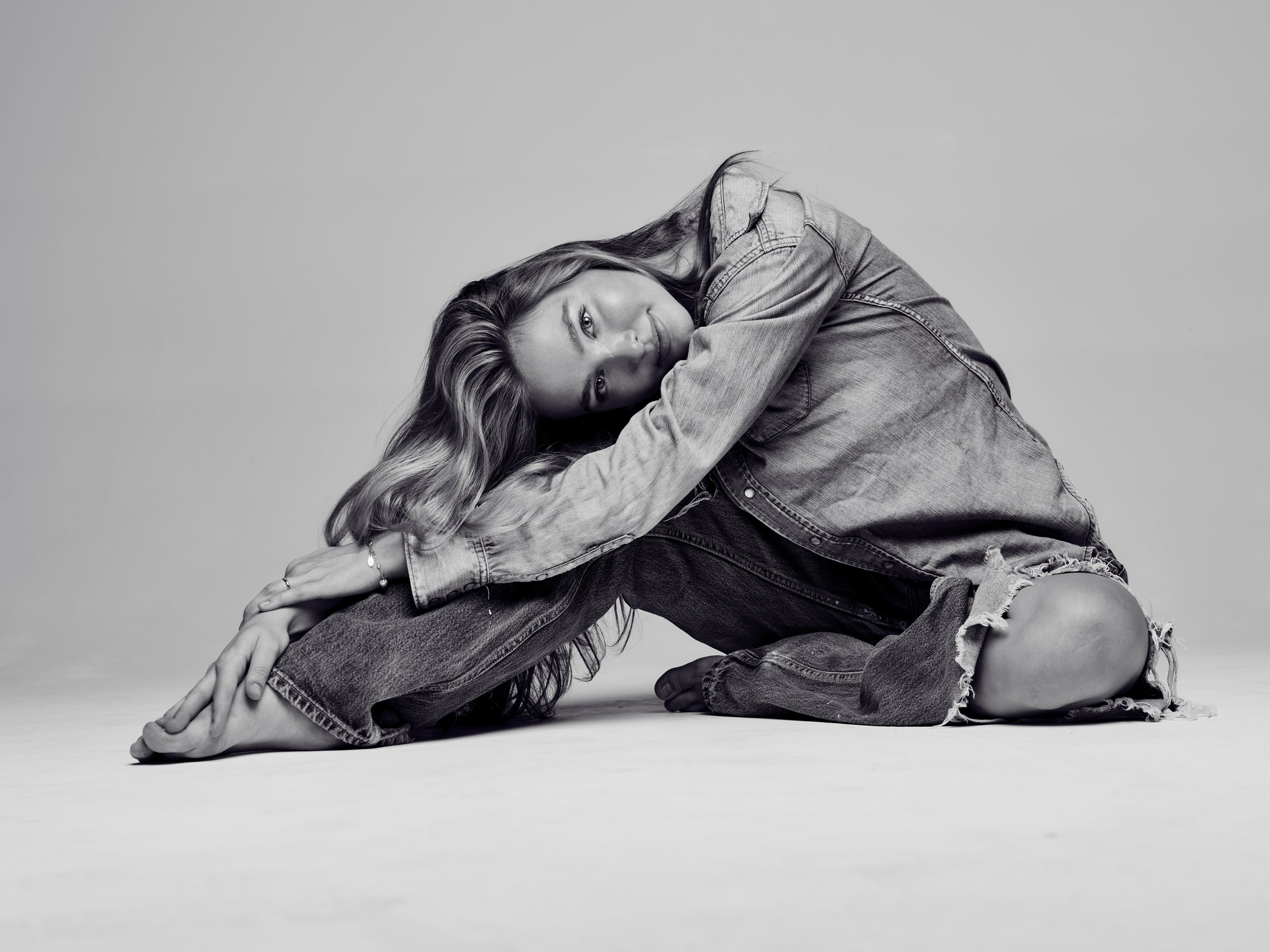 Black and white photo of a woman with long hair wearing a denim jacket and ripped jeans, sitting on the floor with her legs crossed and leaning her head on her arm, smiling slightly.