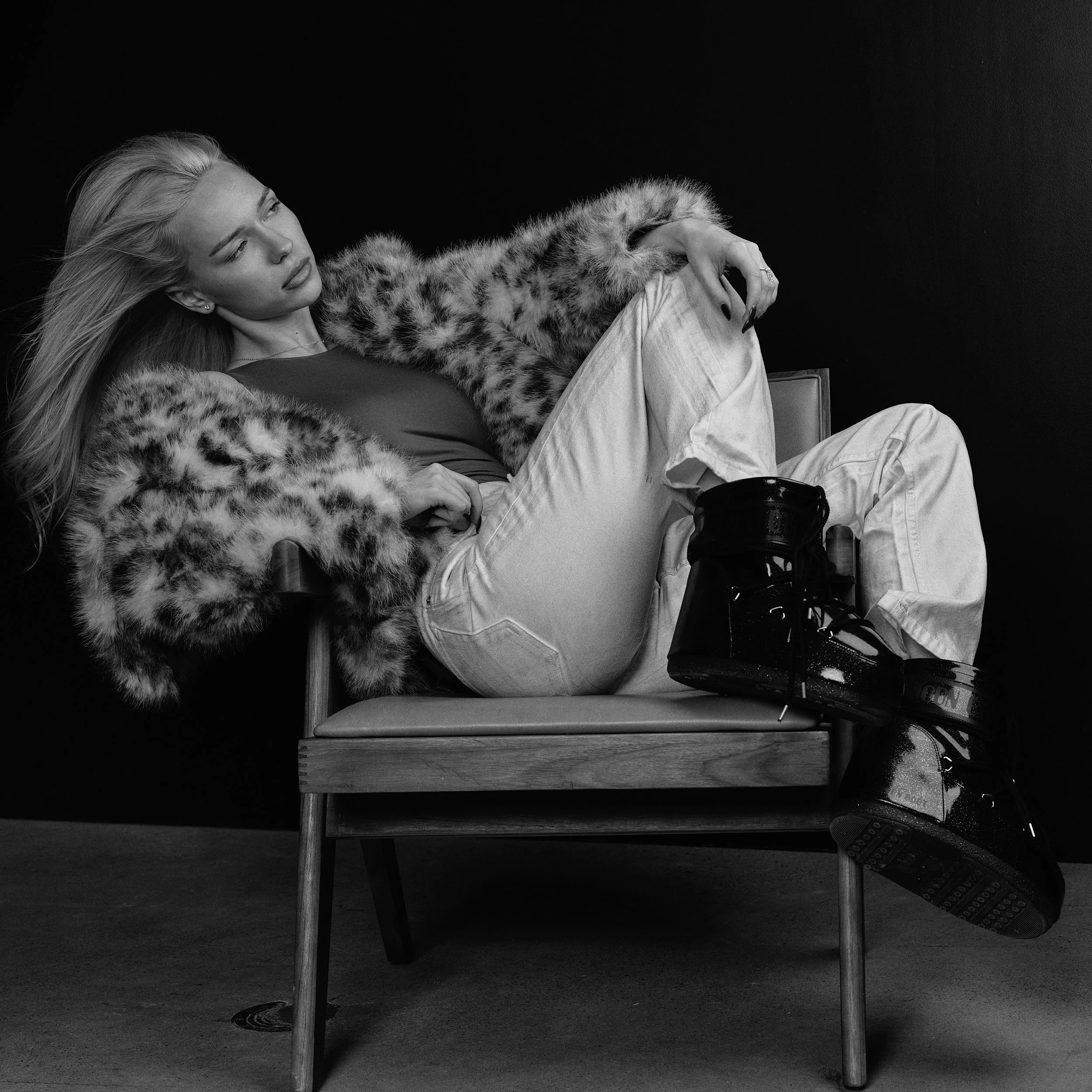Black and white photo of a woman with long hair sitting on a chair with her head leaning on her arm, wearing a fur coat and light-colored pants, against a dark background.