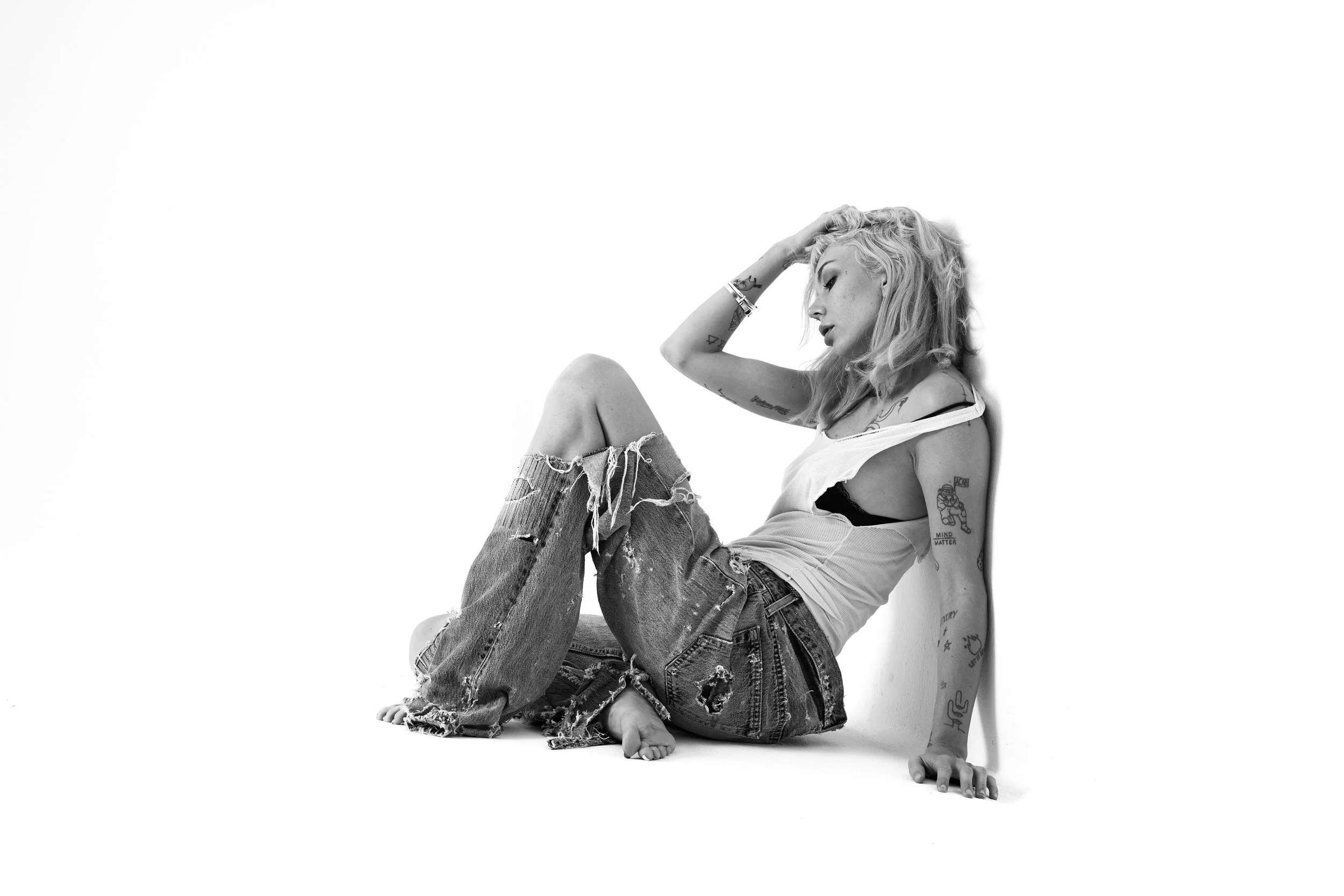 A woman with curly blonde hair, tattoos, and ripped jeans, sitting on the floor with her back against a white wall, resting her head on her hand in a relaxed pose.