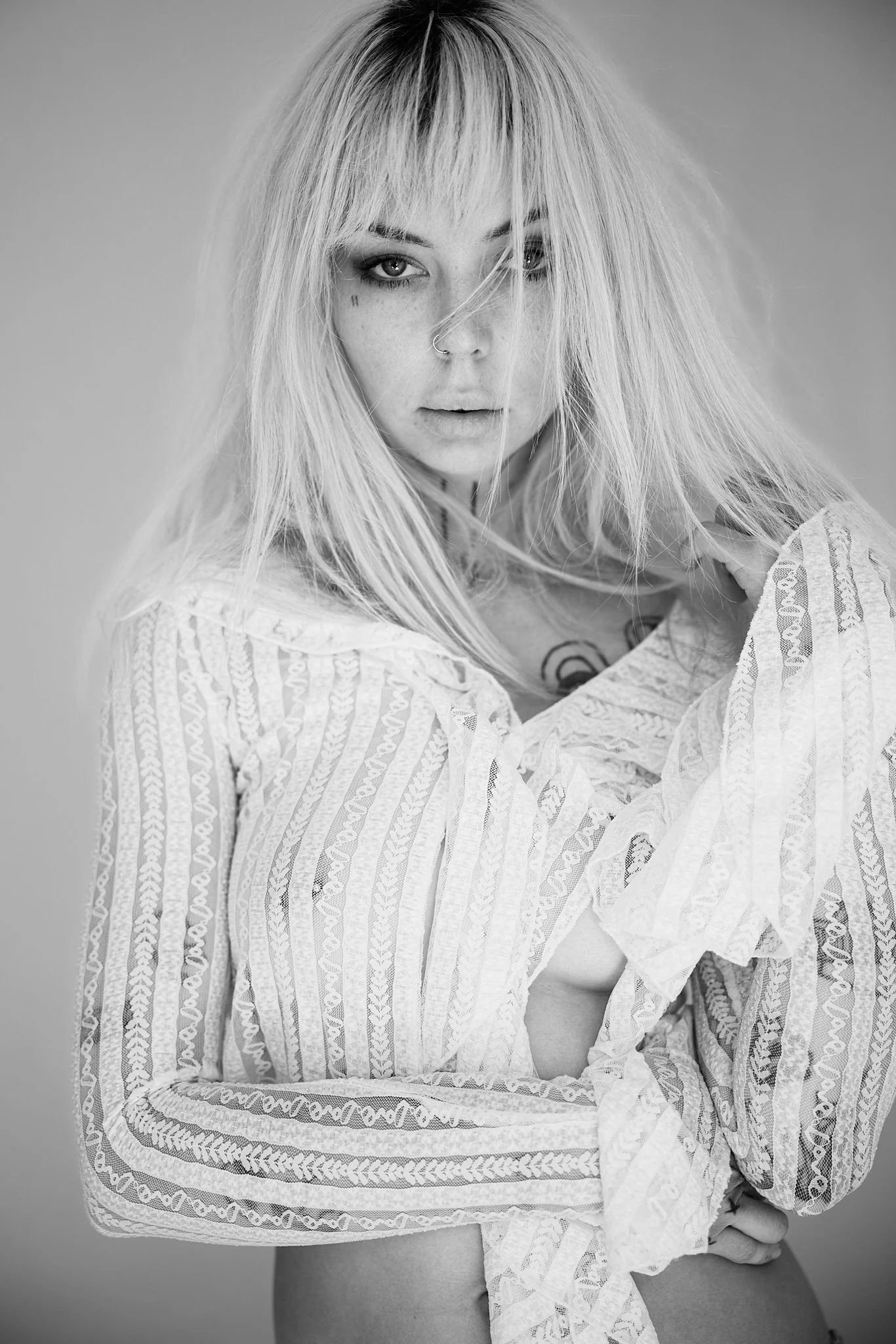 Black and white photo of a woman with long blonde hair, looking at the camera with a serious expression. She is wearing a patterned, loose, long-sleeve top, revealing part of her chest and arm.