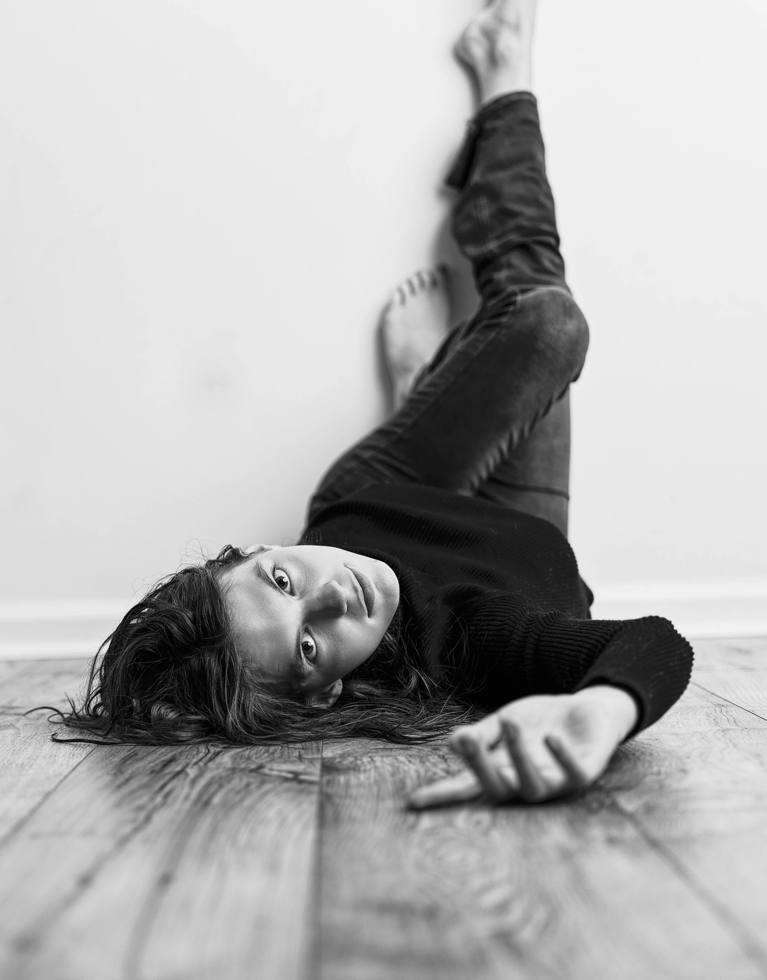 A woman lying on the wooden floor in a black sweater, reaching out with her hand, with her head turned towards the camera and long hair spread on the floor, black and white photo.