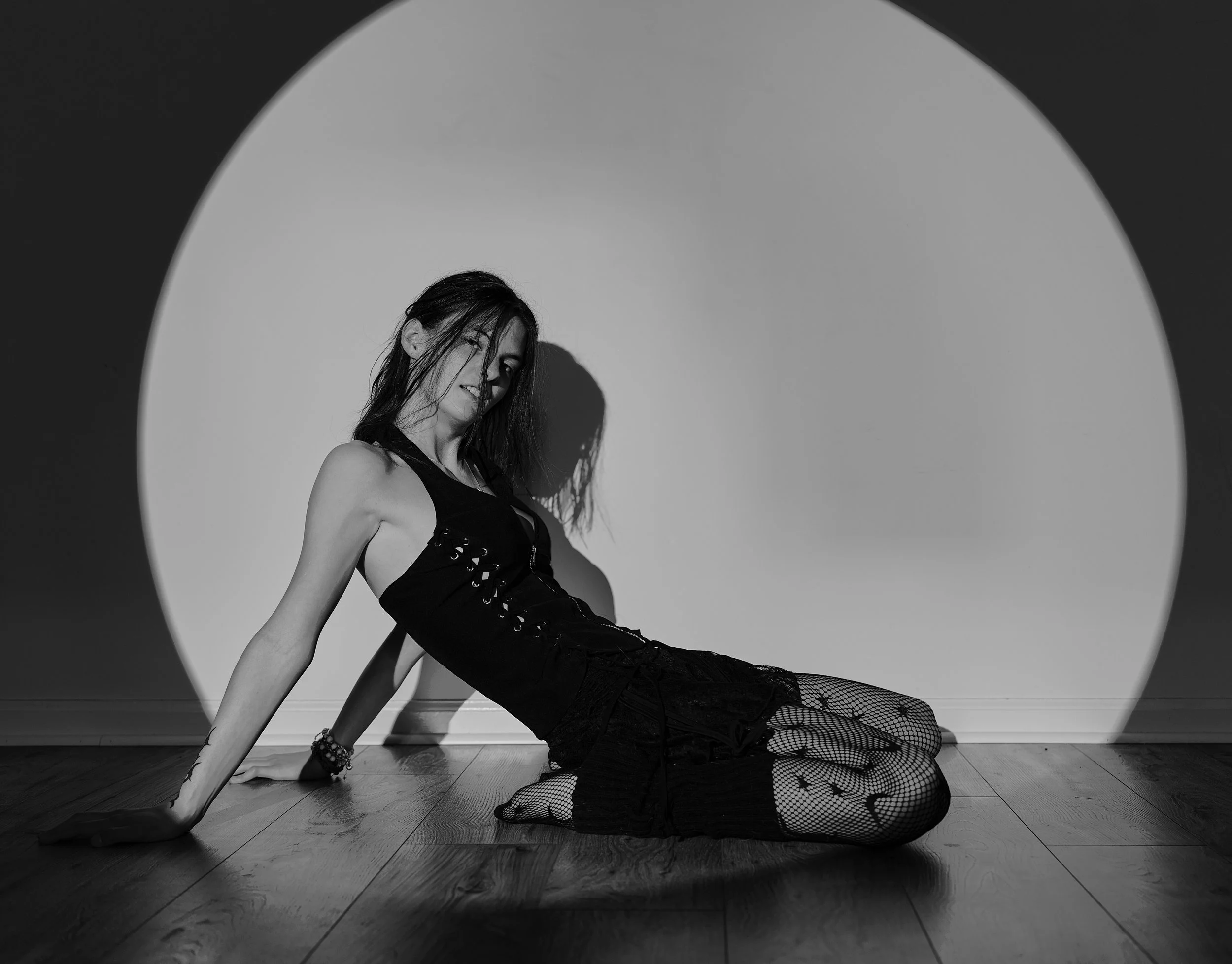 A woman with dark hair posed on the floor against a white background, with her body leaning back and her head tilted to the side, wearing a black outfit and fishnet stockings.