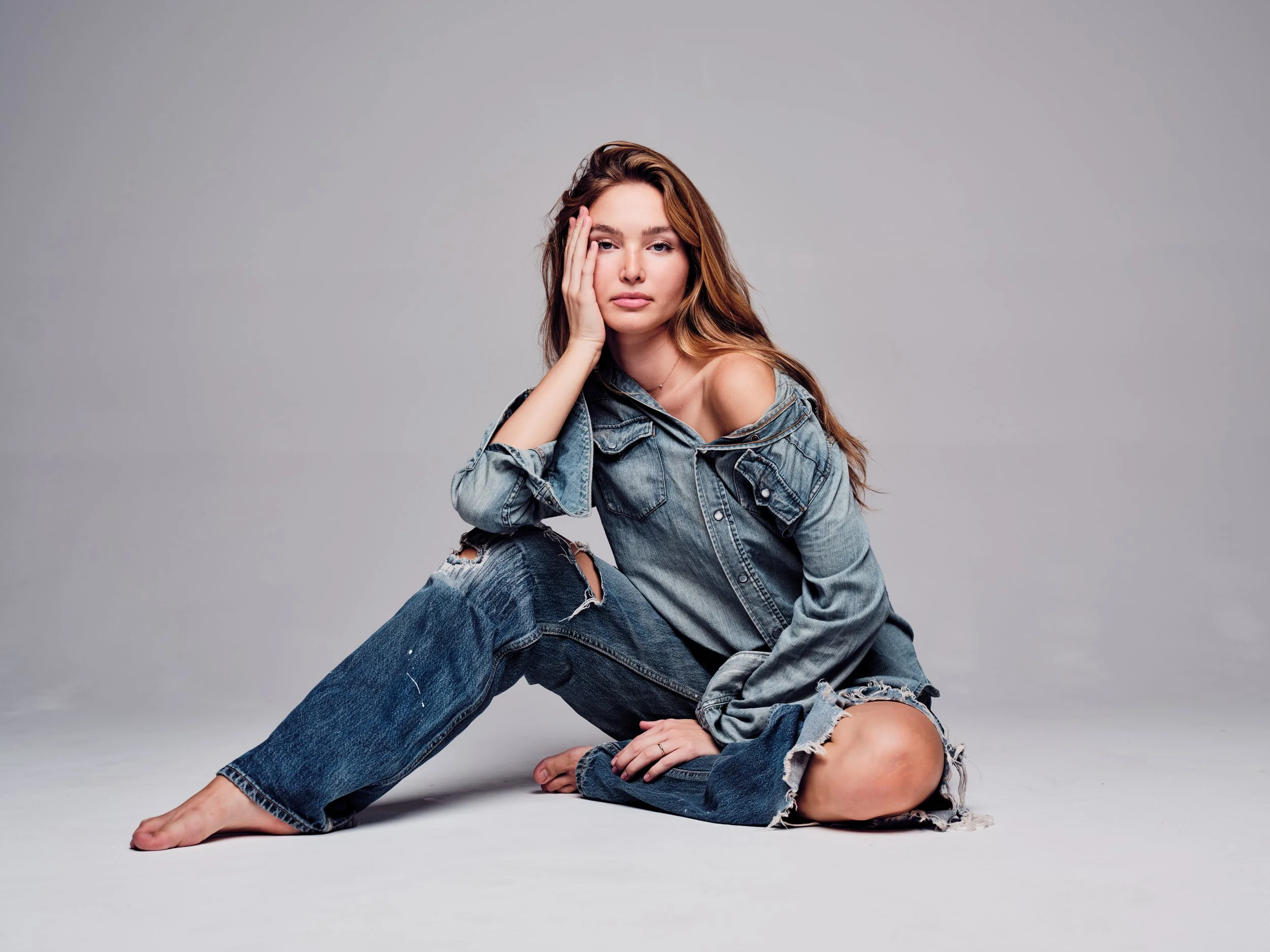 A woman with long brown hair, wearing a ripped denim jacket and jeans, posing on a gray background. She is sitting with one hand on her head and the other arm on her knee, looking at the camera with a neutral expression.