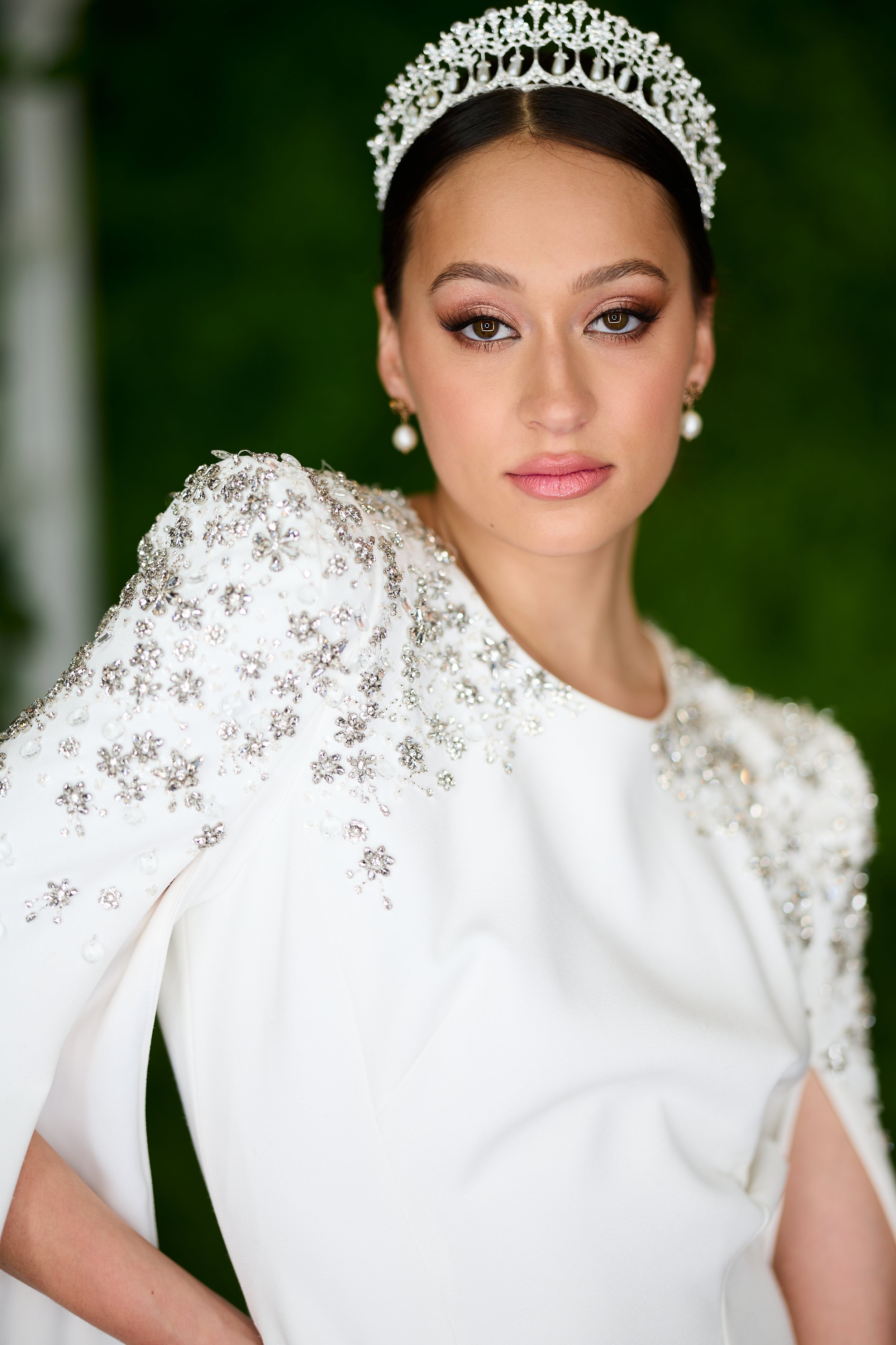 Close-up of a woman in a white dress with silver floral embellishments on the shoulder, wearing pearl earrings and a tiara, with a blurred green background.