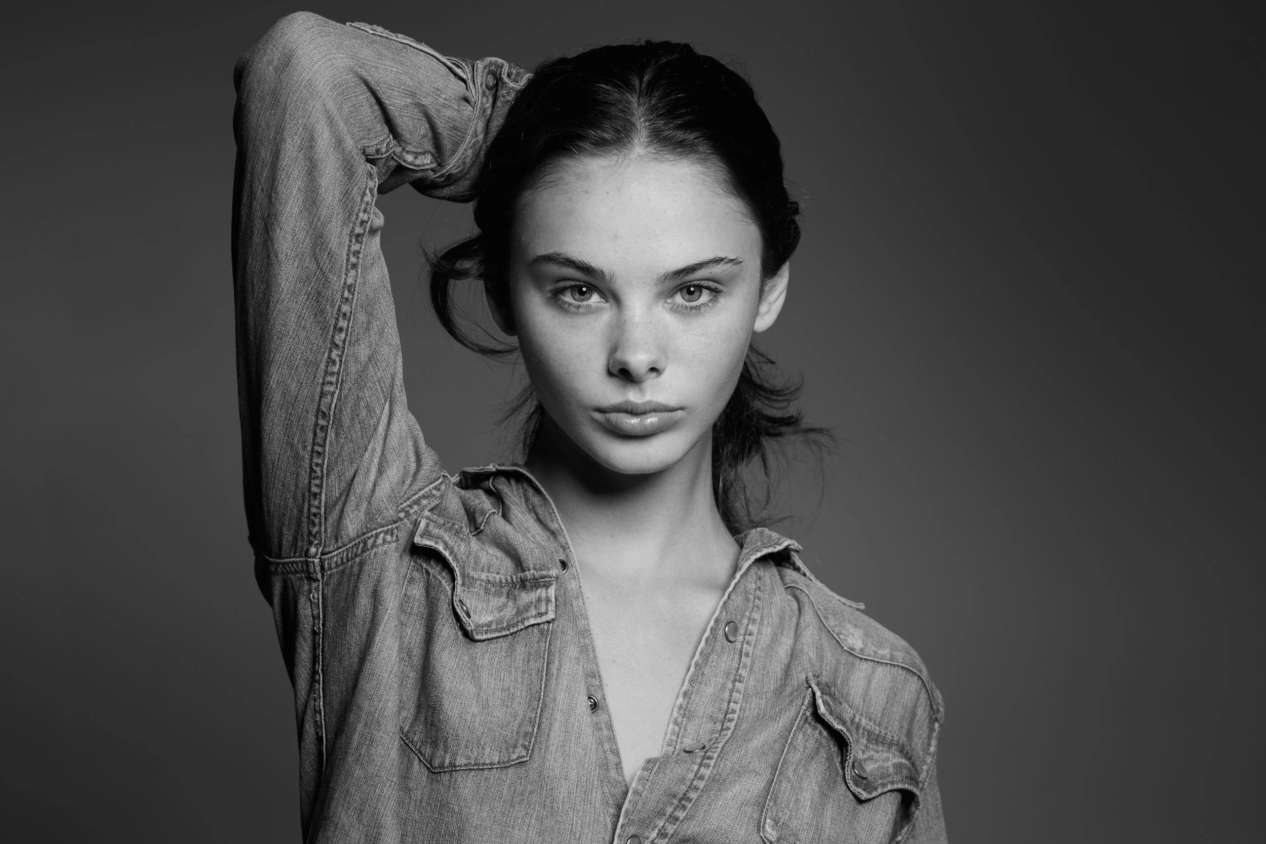 Black and white photo of a young woman with short dark hair, wearing a denim shirt, looking directly at the camera with her hand resting behind her head.