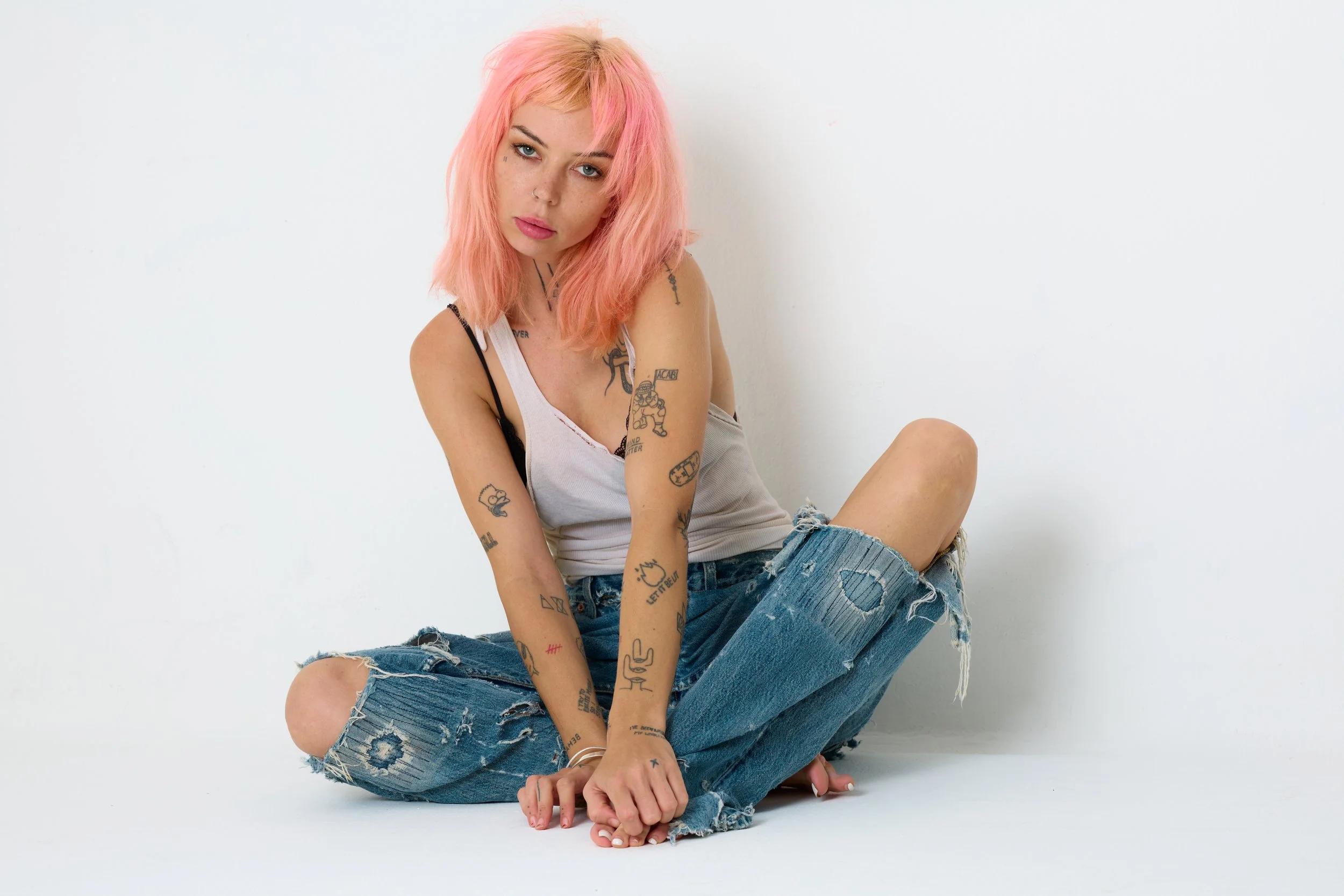 A young woman with pink hair and tattoos sitting on the floor against a white background.