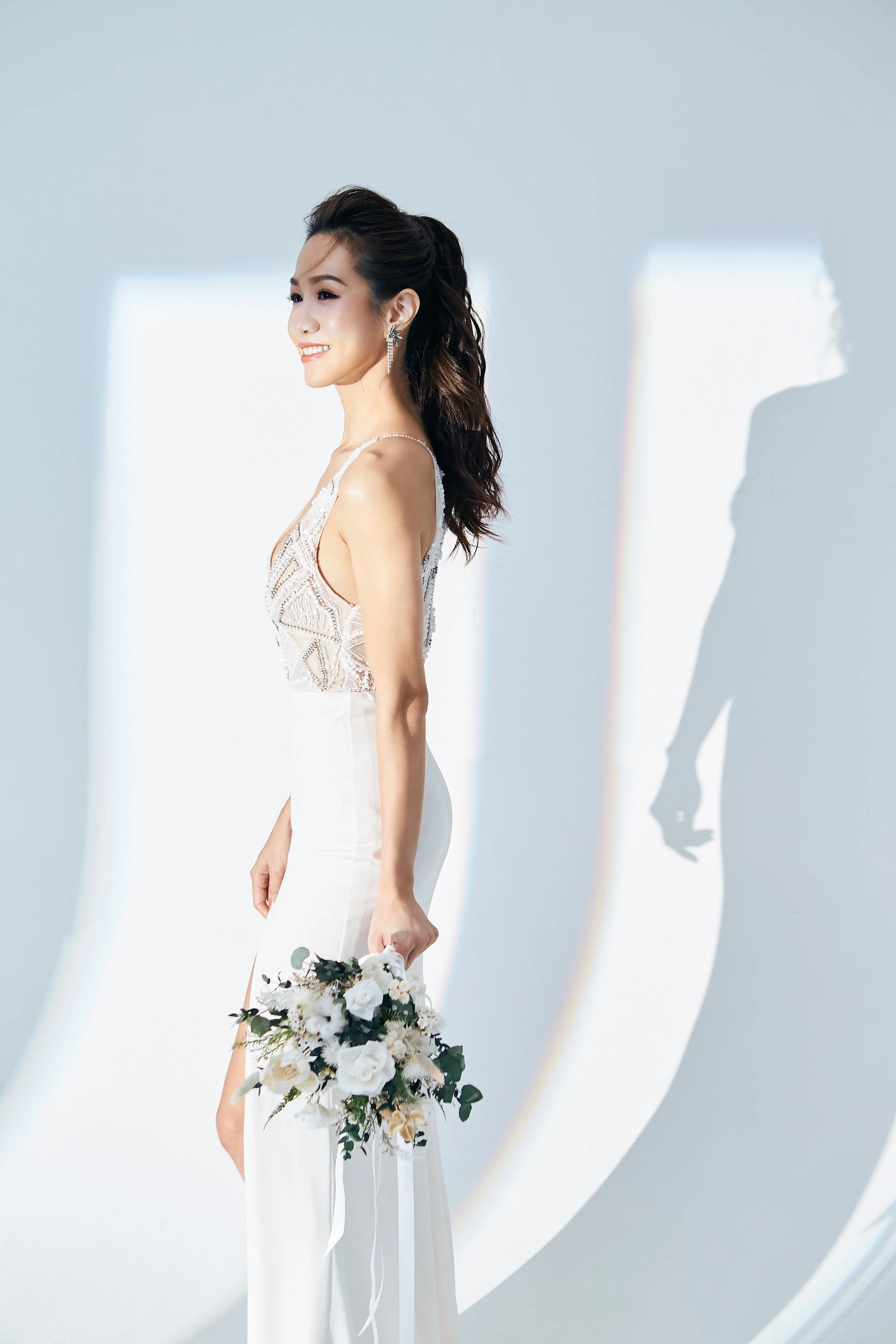 A bride in a white wedding dress is holding a bouquet of white flowers and greenery, standing against a light blue background with sunlight creating her shadow on the wall.