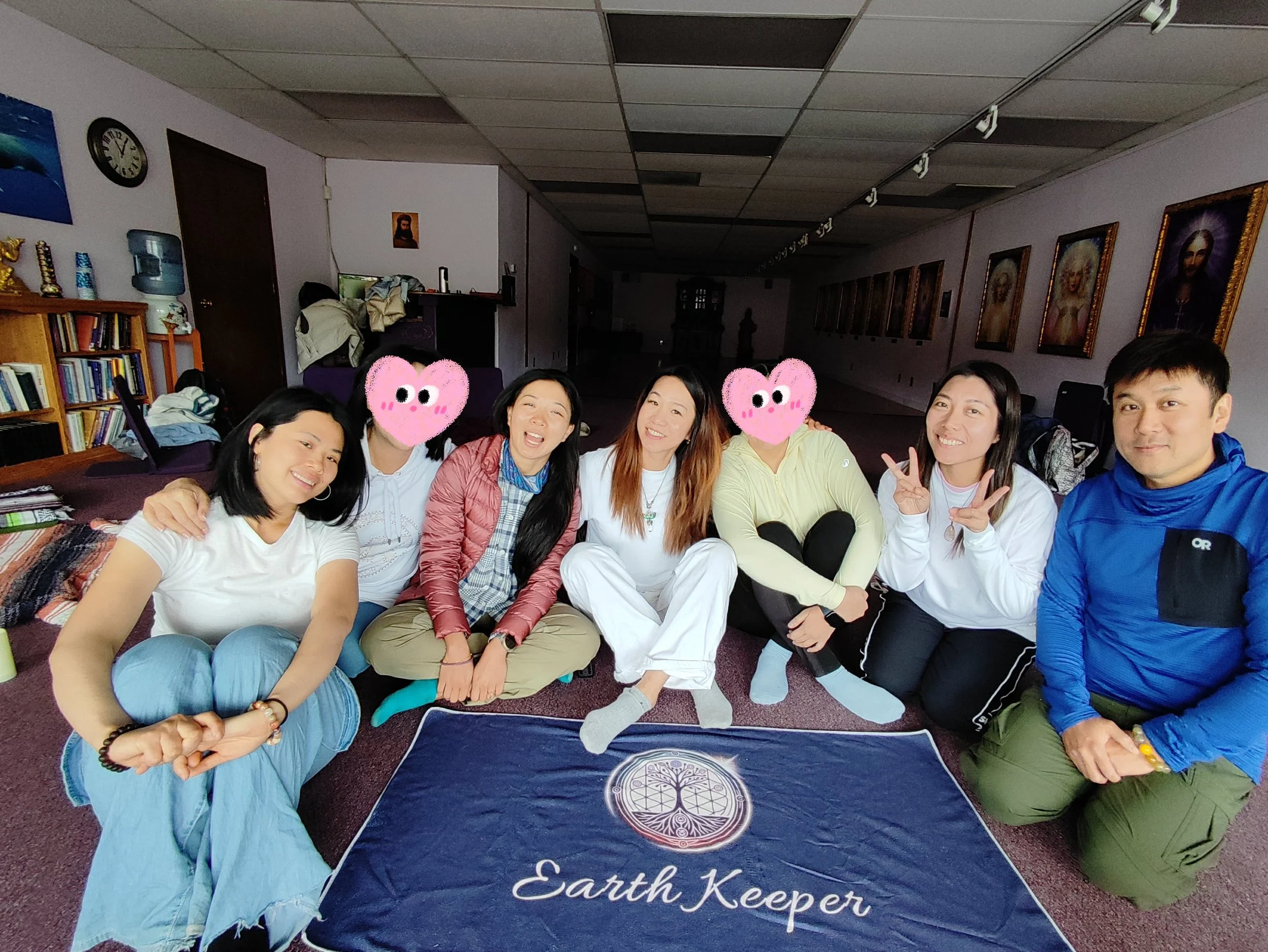 A group of seven people sitting on the floor inside a room, smiling at the camera, with paintings on the wall and a bookshelf in the background. The two individuals with hearts replacing their faces are in the middle. A cloth with a tree and Earth Ke