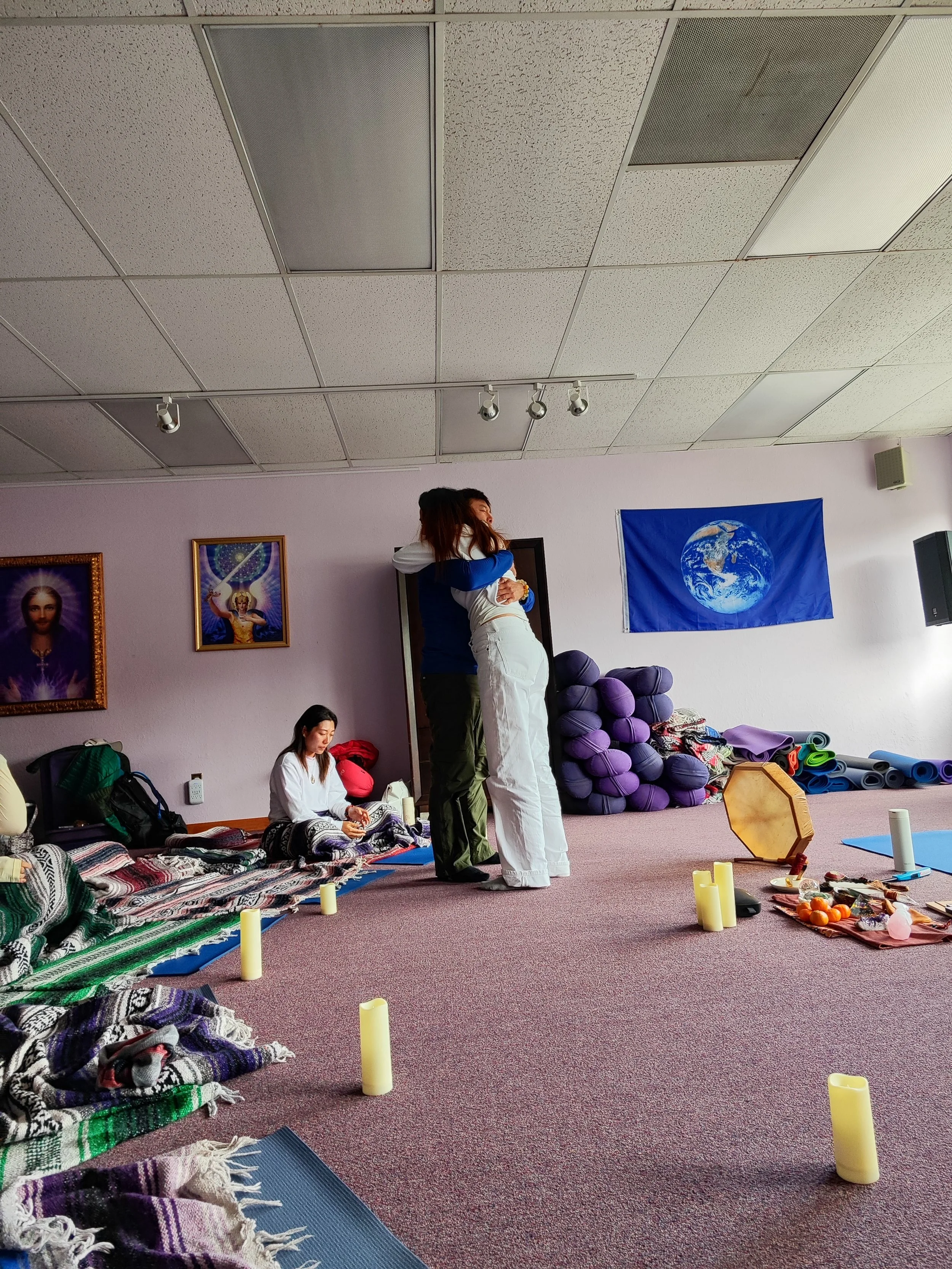 People participating in a yoga or meditation class in a room decorated with spiritual posters, a globe banner, and various yoga props, with candles placed on the floor.