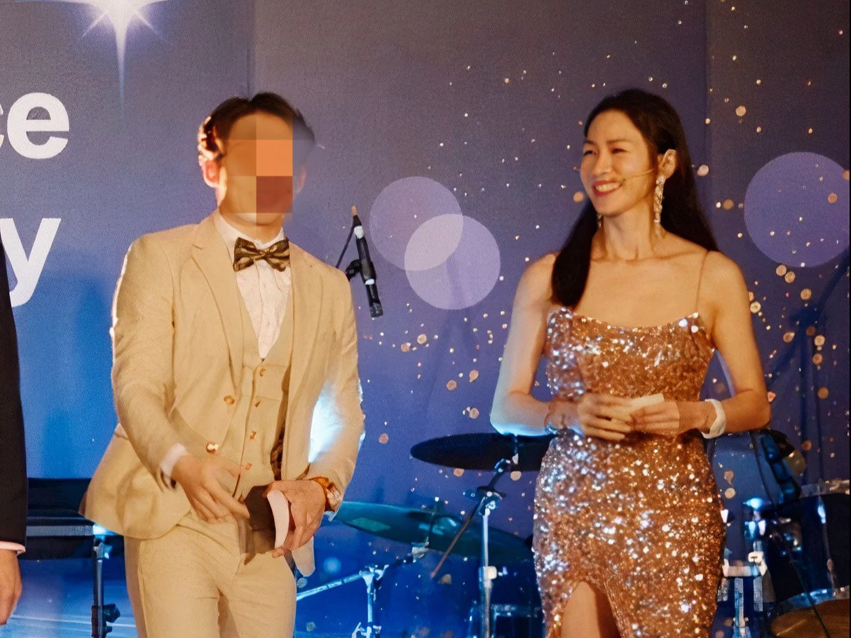 A man in a cream-colored suit with a bow tie and a woman in a shiny, gold sequin dress standing on stage at an event, with a dark background and musical instruments behind them.