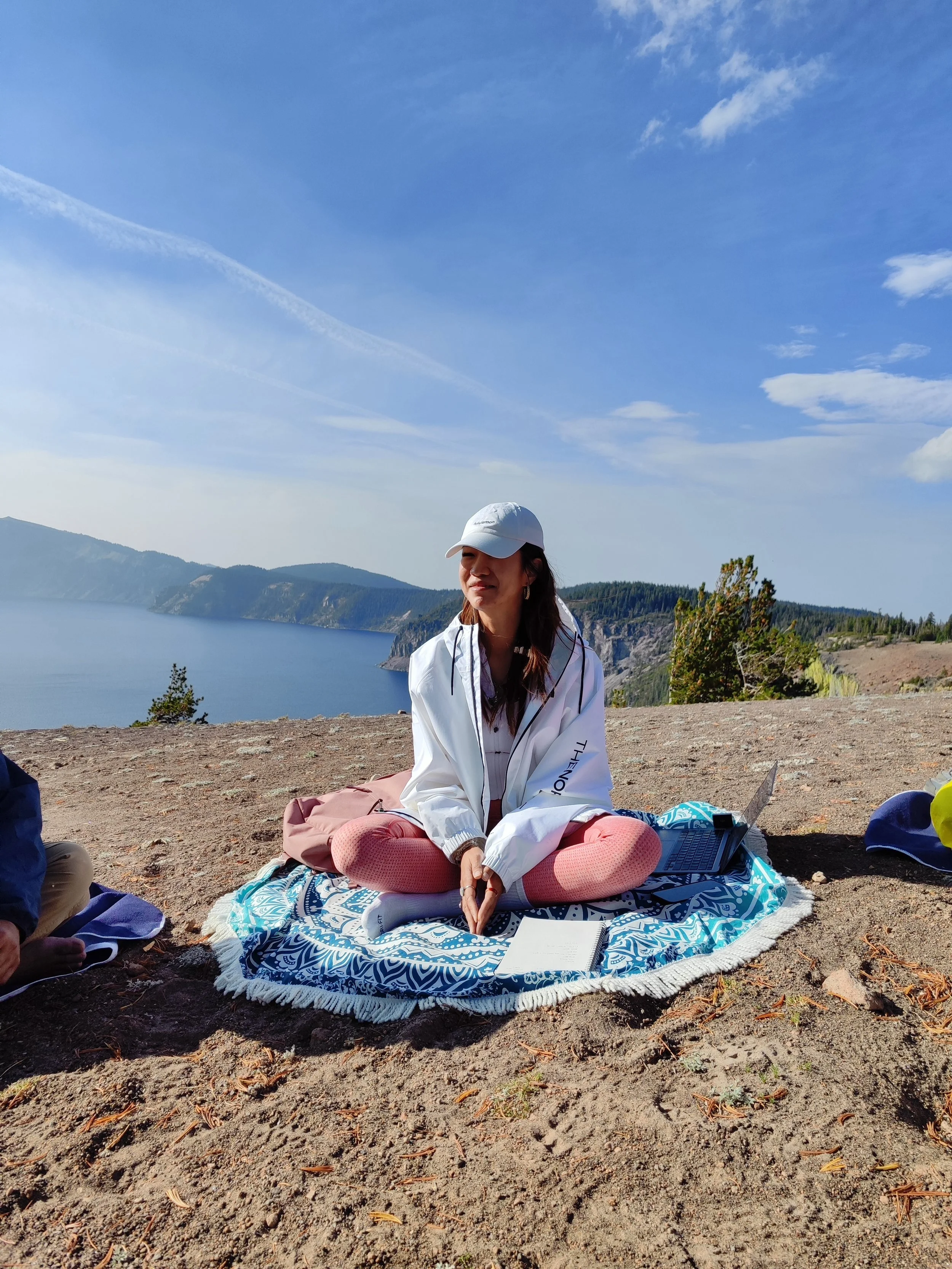 A woman sitting cross-legged on an outdoor blanket, wearing a white cap and white jacket, with a laptop and notebook in front of her. The background features a large lake, hills, and a blue sky with scattered clouds.