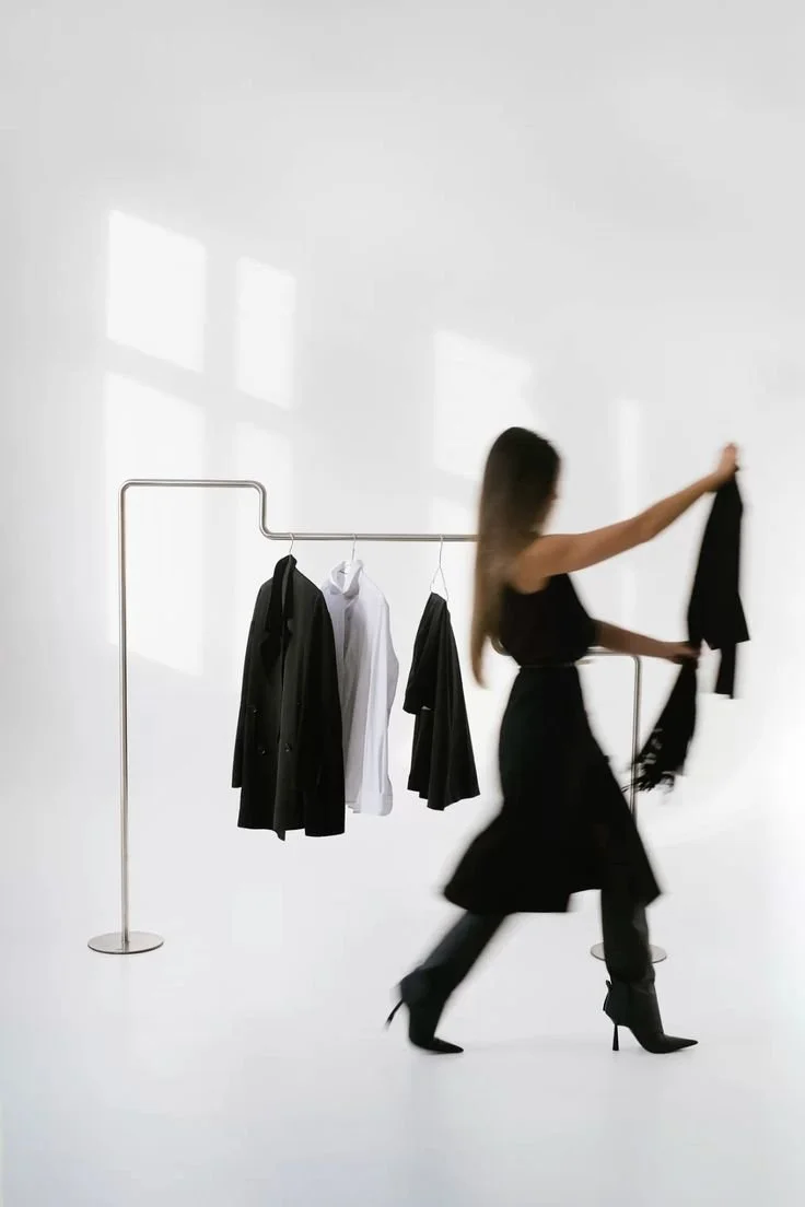 A woman in high heels browsing black clothing on a rack in a minimal, bright room with white walls and floor.