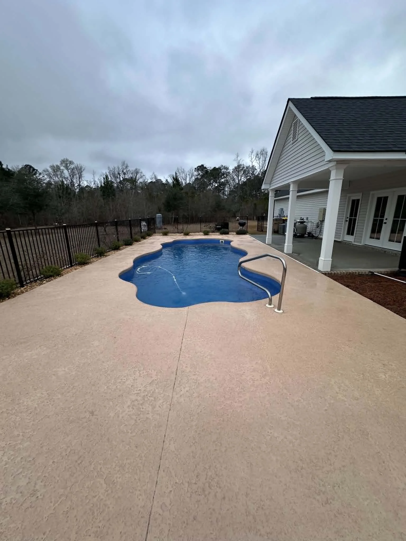 Residential backyard patio with an in-ground kidney-shaped swimming pool, surrounded by a black metal fence, with outdoor grill and storage area under a covered porch, and a gray cloudy sky overhead.