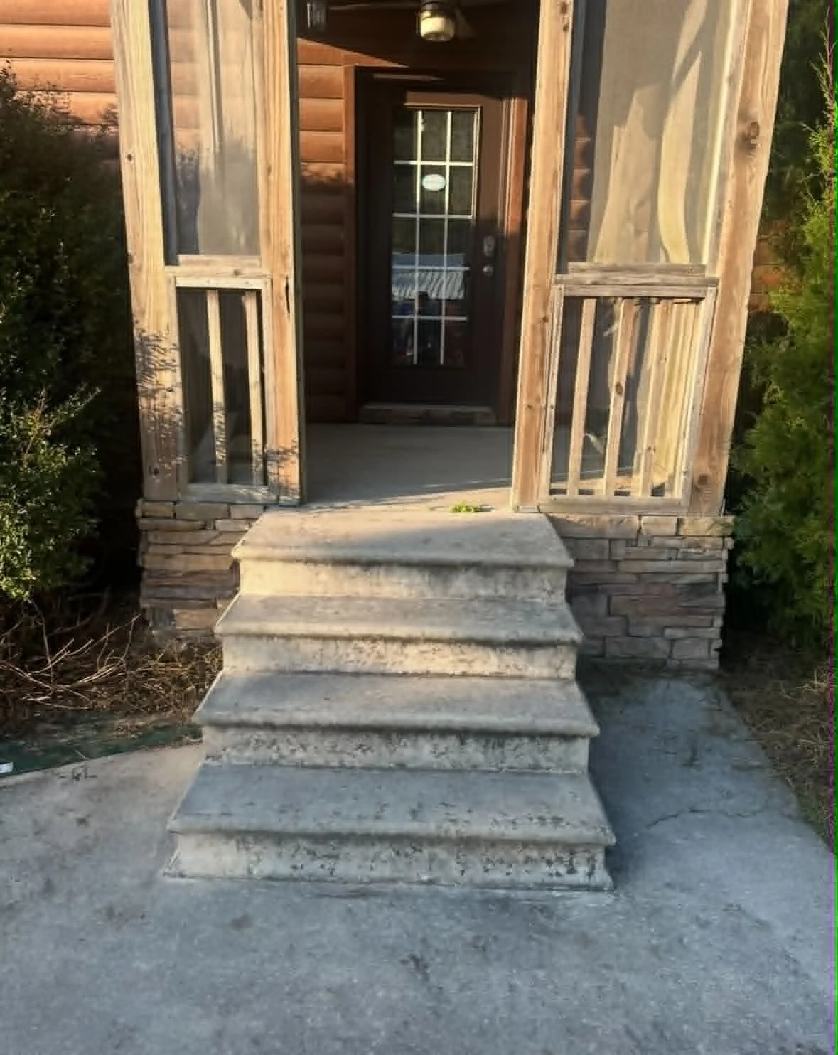 Front view of a small porch with three concrete steps leading to a wooden door with glass panes, surrounded by wooden and brick siding, and green plants on the sides.
