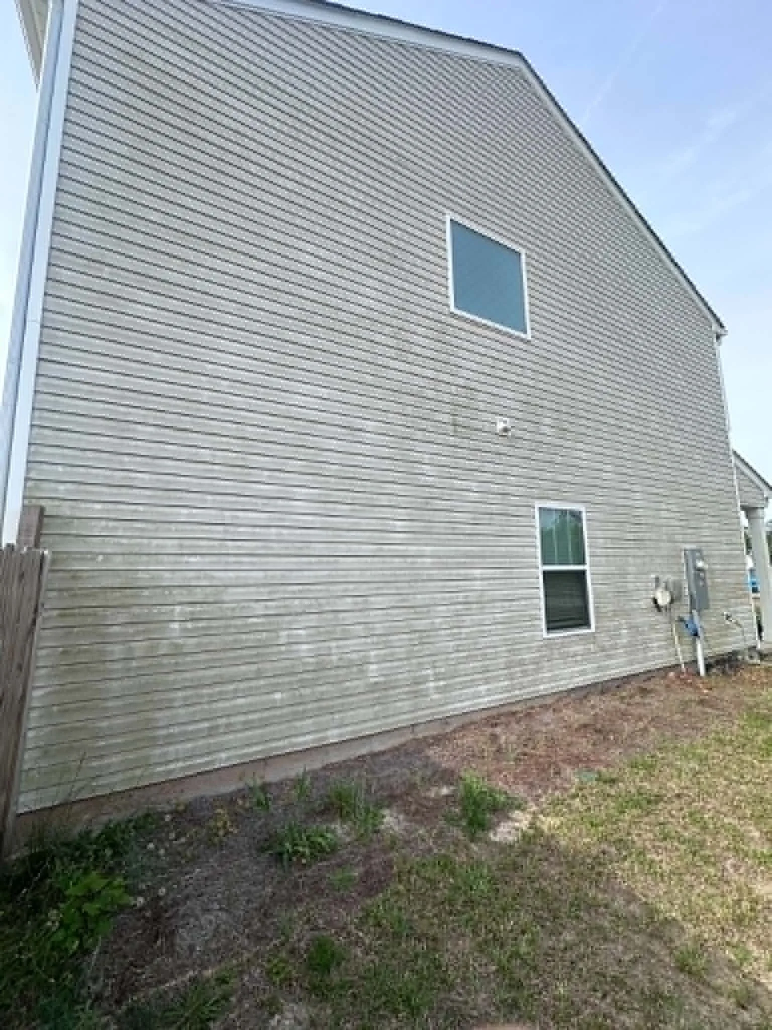 Rear view of a house with beige vinyl siding, two small windows, and a small outdoor utility area with pipes and electrical meters, surrounded by grass and dirt.