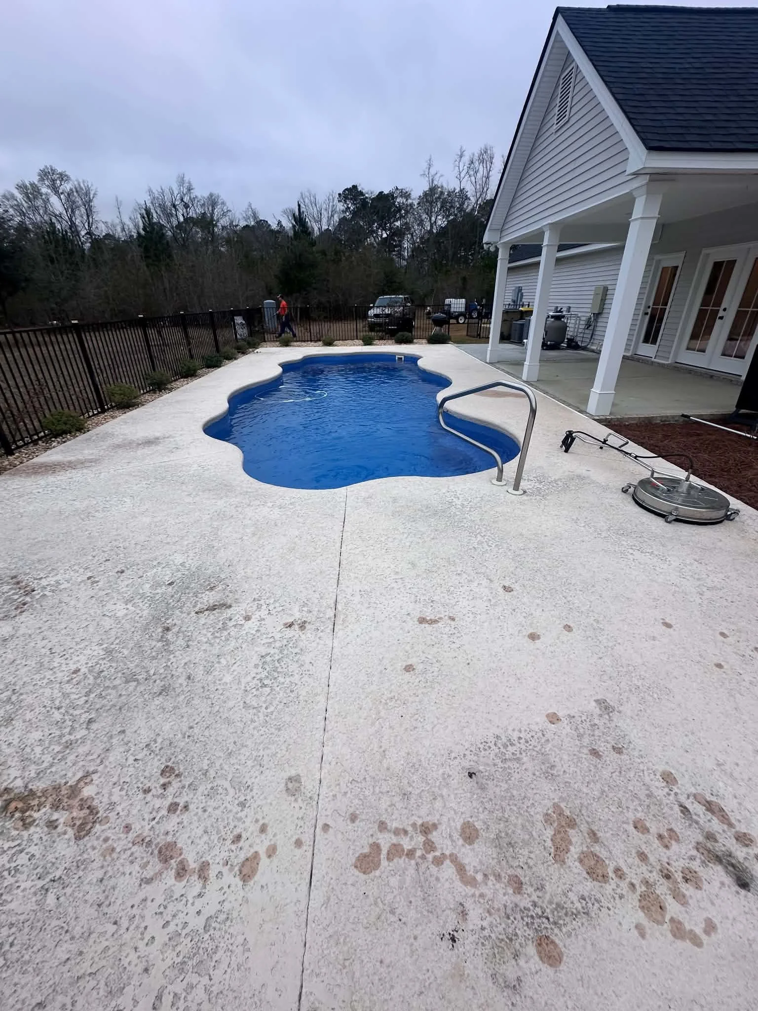 Backyard with in-ground swimming pool, white concrete deck, house with covered patio, outdoor furniture and equipment, trees in the background, cloudy sky.