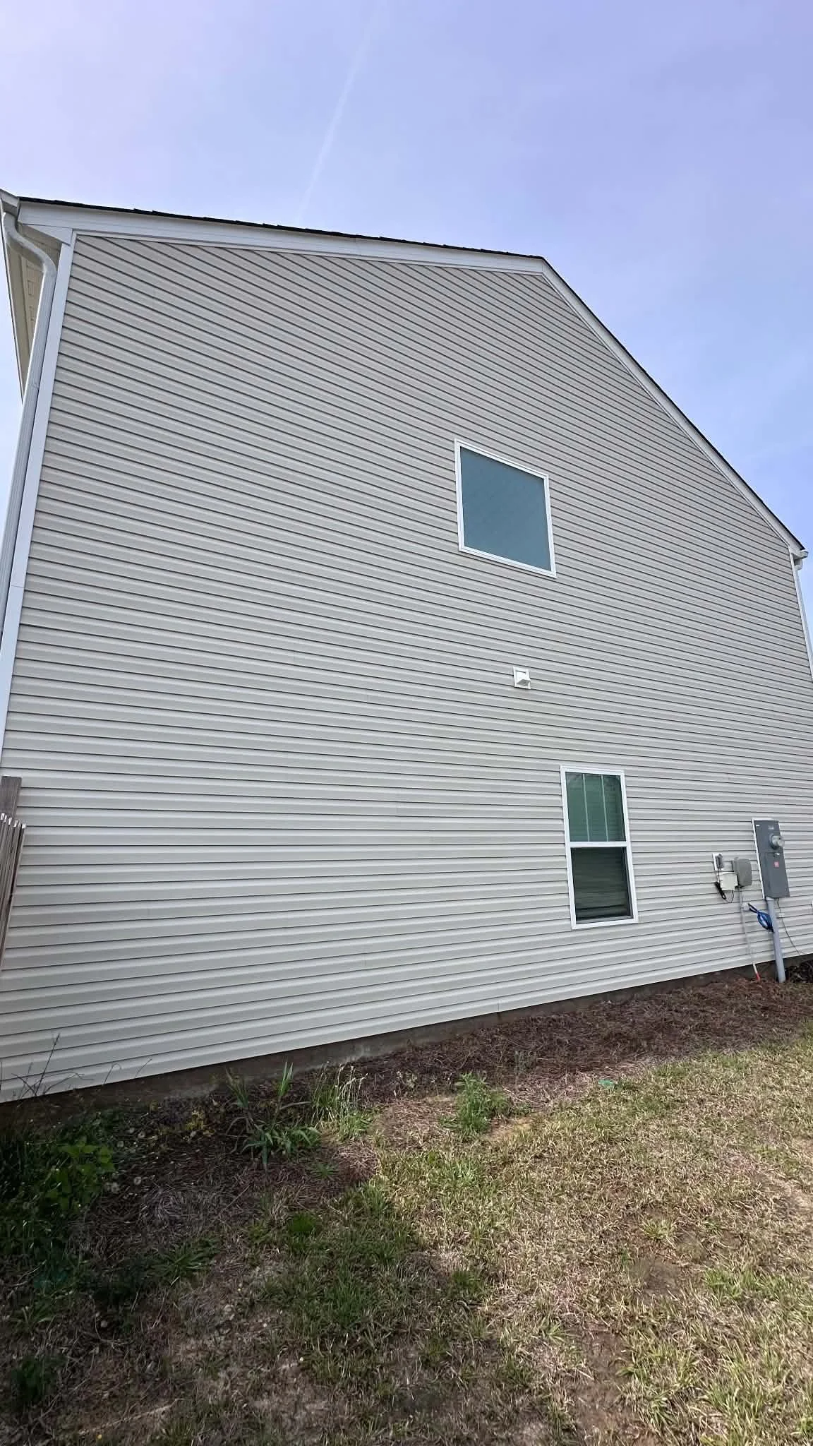The side of a house with beige vinyl siding, two windows, an electrical box, and outdoor utility meters. The ground is bare soil and grass.
