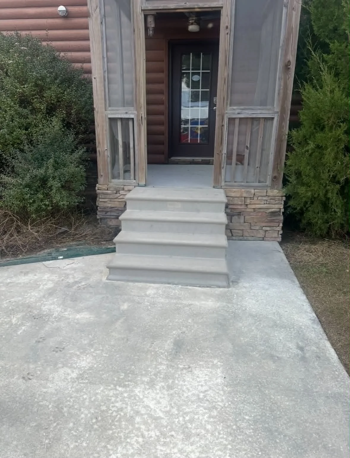 Front entrance of a house with a small staircase leading up to a door. The house has a wooden exterior with a brick foundation, and there are bushes on either side of the entrance.