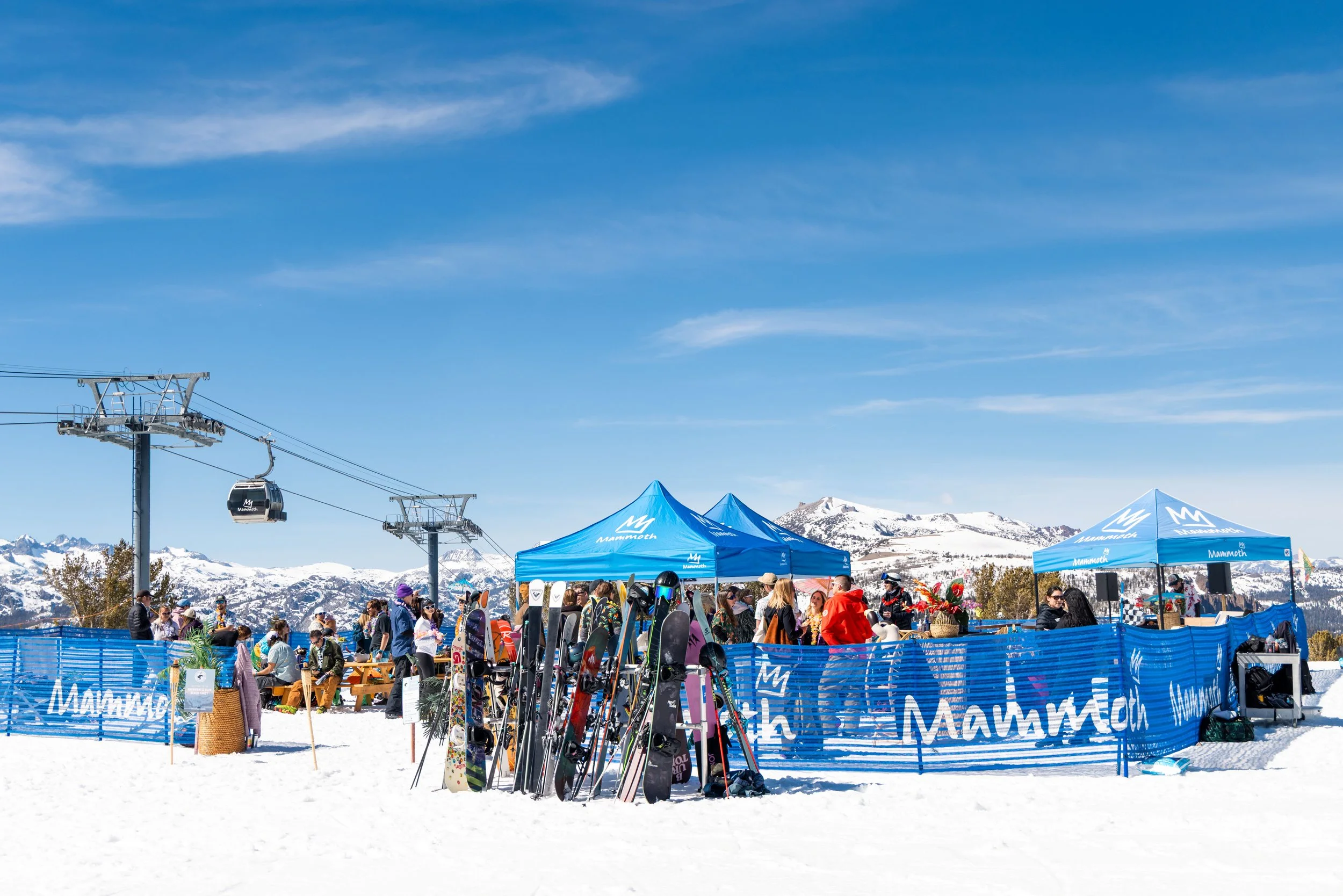 A ski resort scene with ski lifts, blue tents, and people gathered in snow, mountains in the background, and a clear blue sky. Mammoth lakes event for  community foundation donor day. featuring smores, skiing, lunch, dj and, sun bum.
