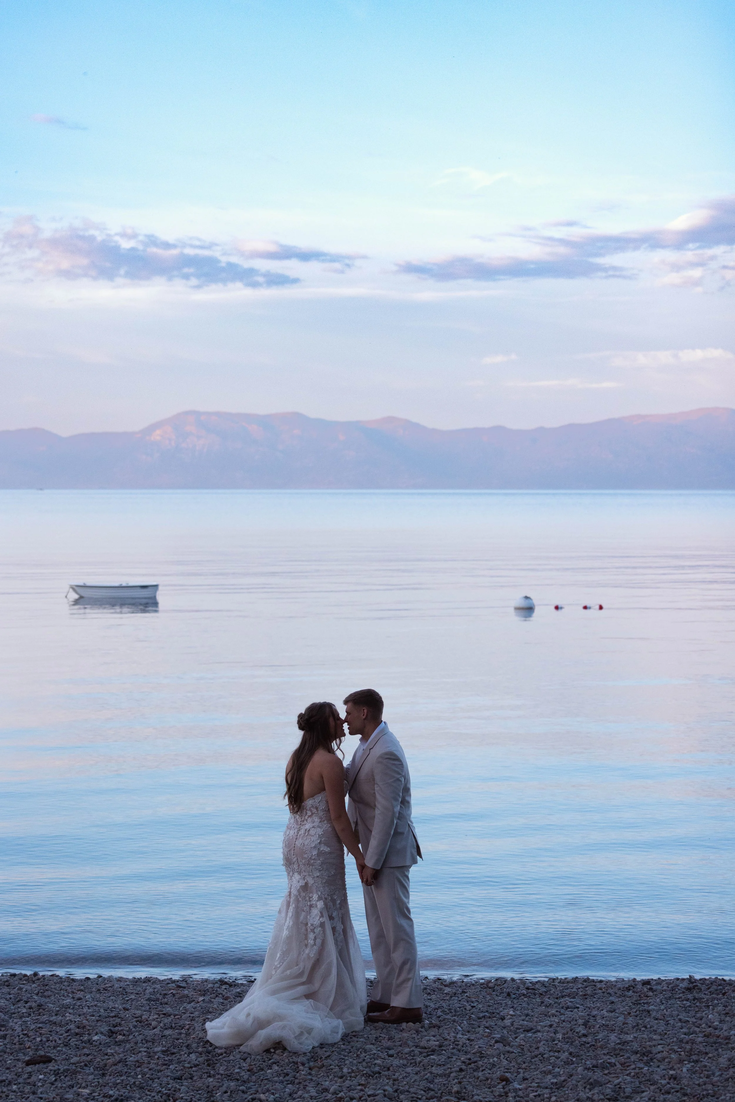 A bride and groom standing by the water's edge, holding hands, with mountains in the background and a boat floating on the water. Lake Tahoe Wedding photos at sunset after they got married at their family's house. 