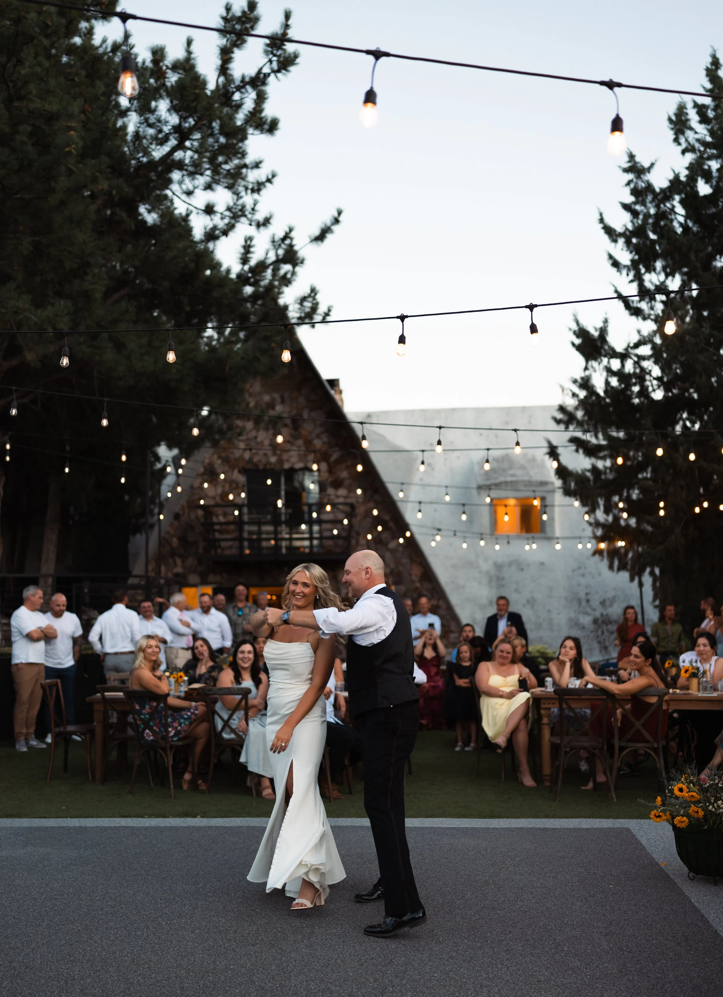 A bride and groom sharing their first dance at an outdoor wedding reception during sunset, with guests seated and standing around them and string lights overhead. stone crossing, eagle Idaho, wedding, in the summer, august. 