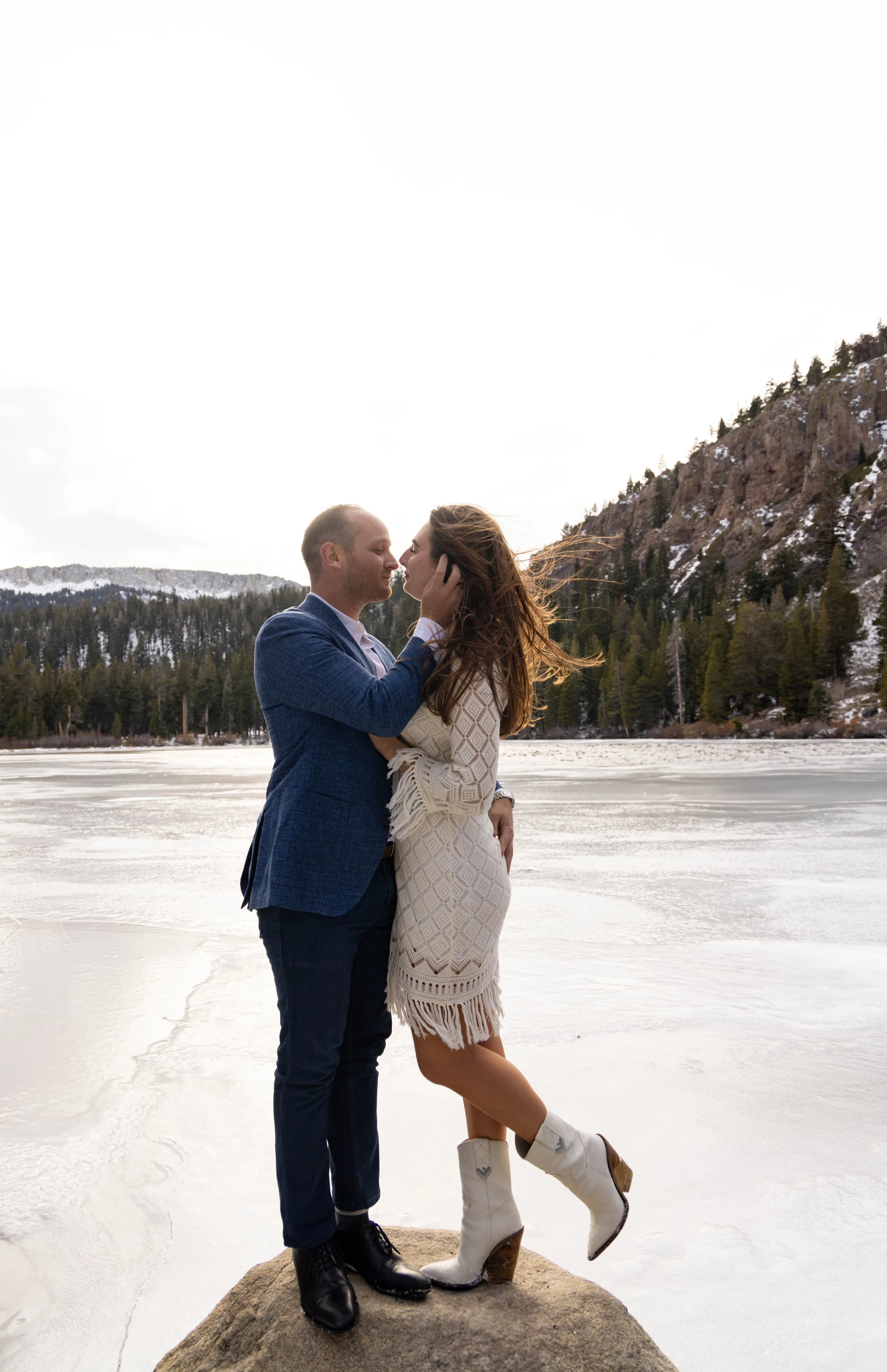 A couple stands on a large rock in a snowy landscape, embracing and staring into each other's eyes, with snow-covered ground and mountains with trees in the background. Twin lakes, engagement photoshoot in mammoth lakes California. 