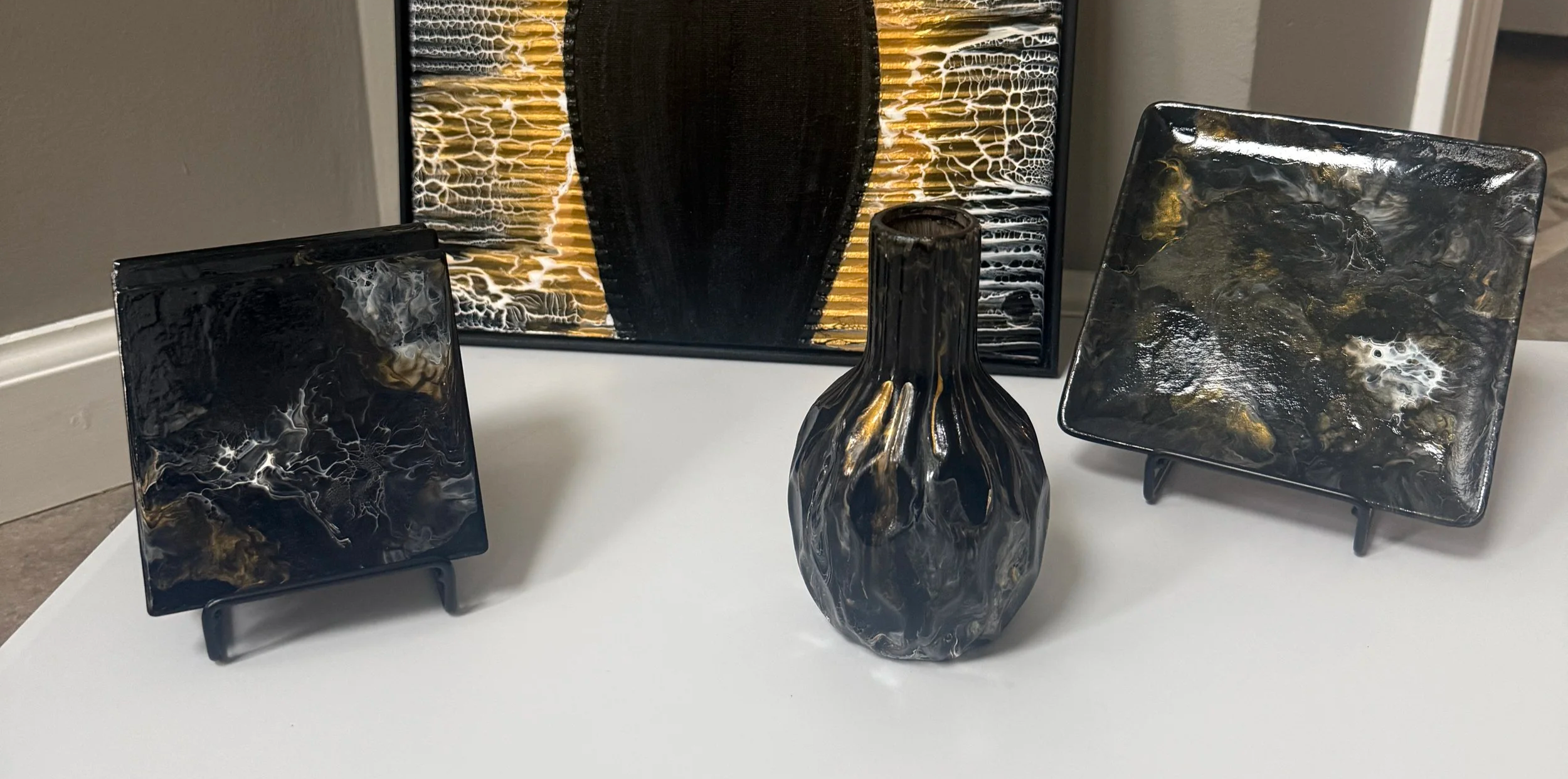 Collection of original abstract fluid art and functional décor pieces in black, gold, white, and charcoal, showcasing coordinated wall art, ceramics, and decorative trays.