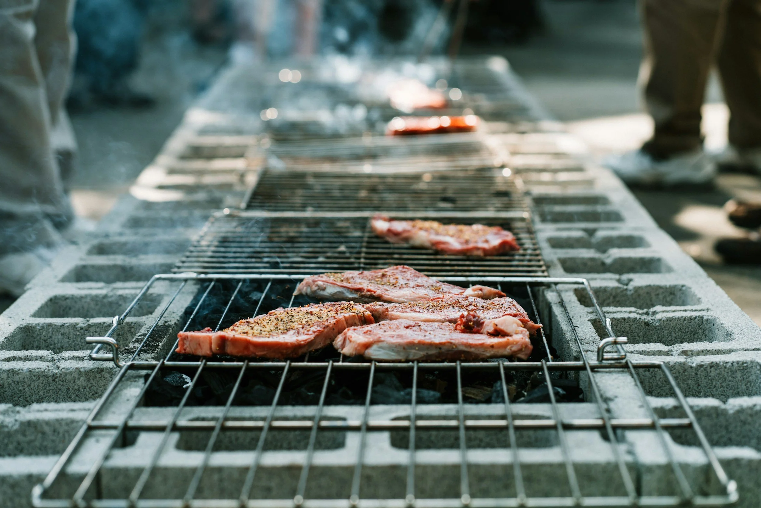 Raw steaks cooking on a grill over an open flame, with some smoke visible in the background.
