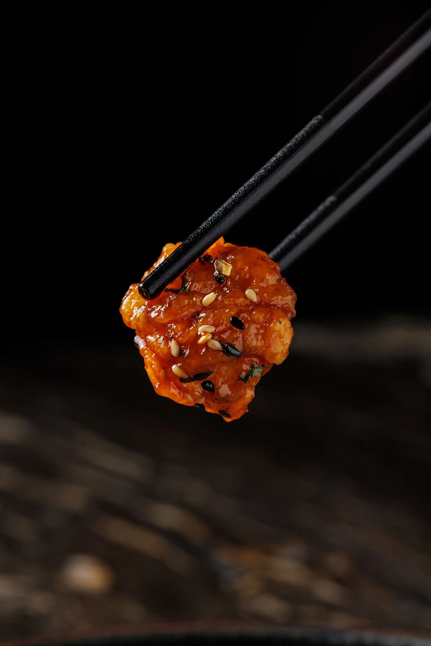 Close-up of a piece of orange glazed meat with sesame seeds, being held with black chopsticks against a black background.