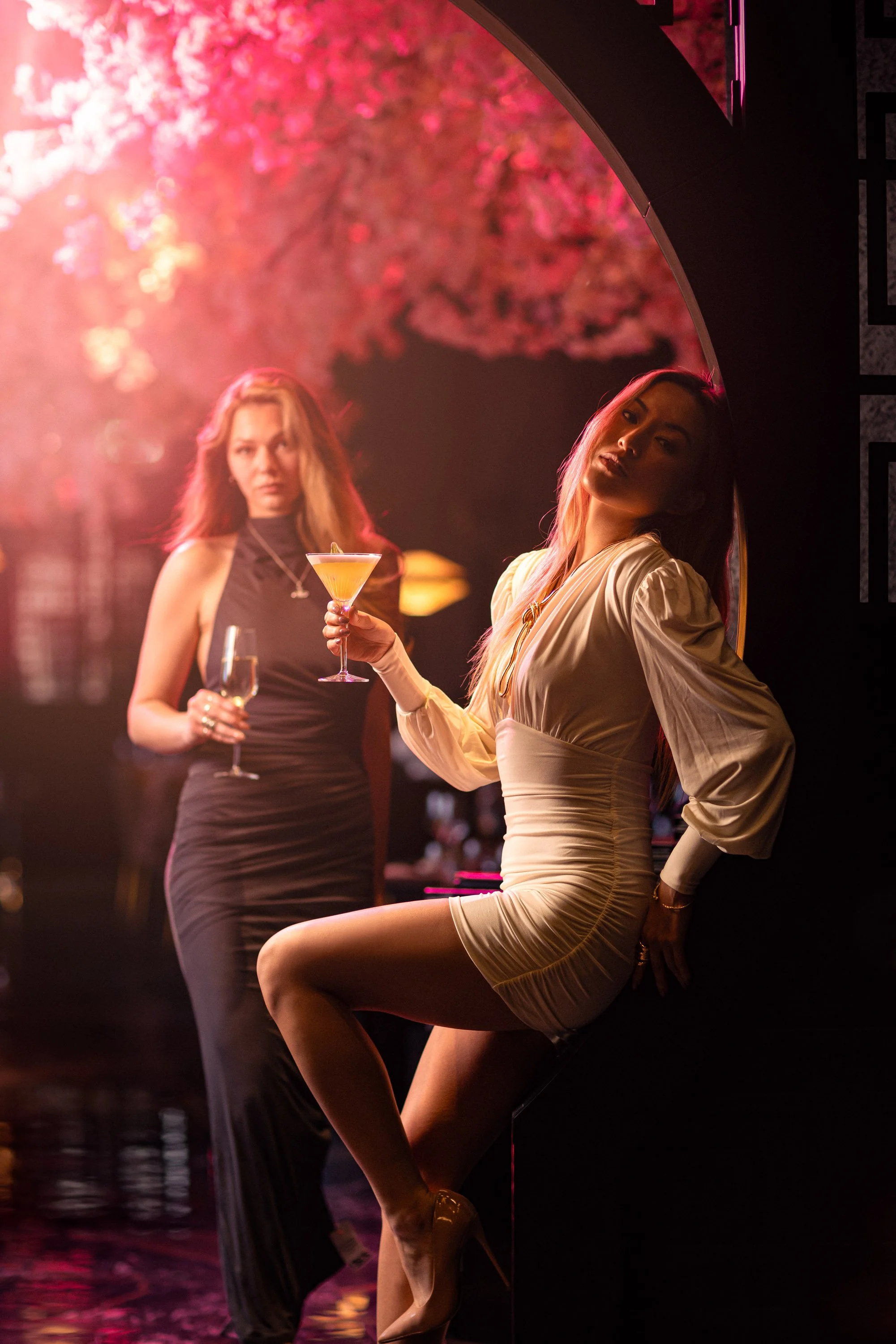Two women at a bar or nightclub; one sitting on a ledge in a beige dress and heels, the other standing in a black dress holding a cocktail. Pink artificial flower decorations and dim lighting are in the background.
