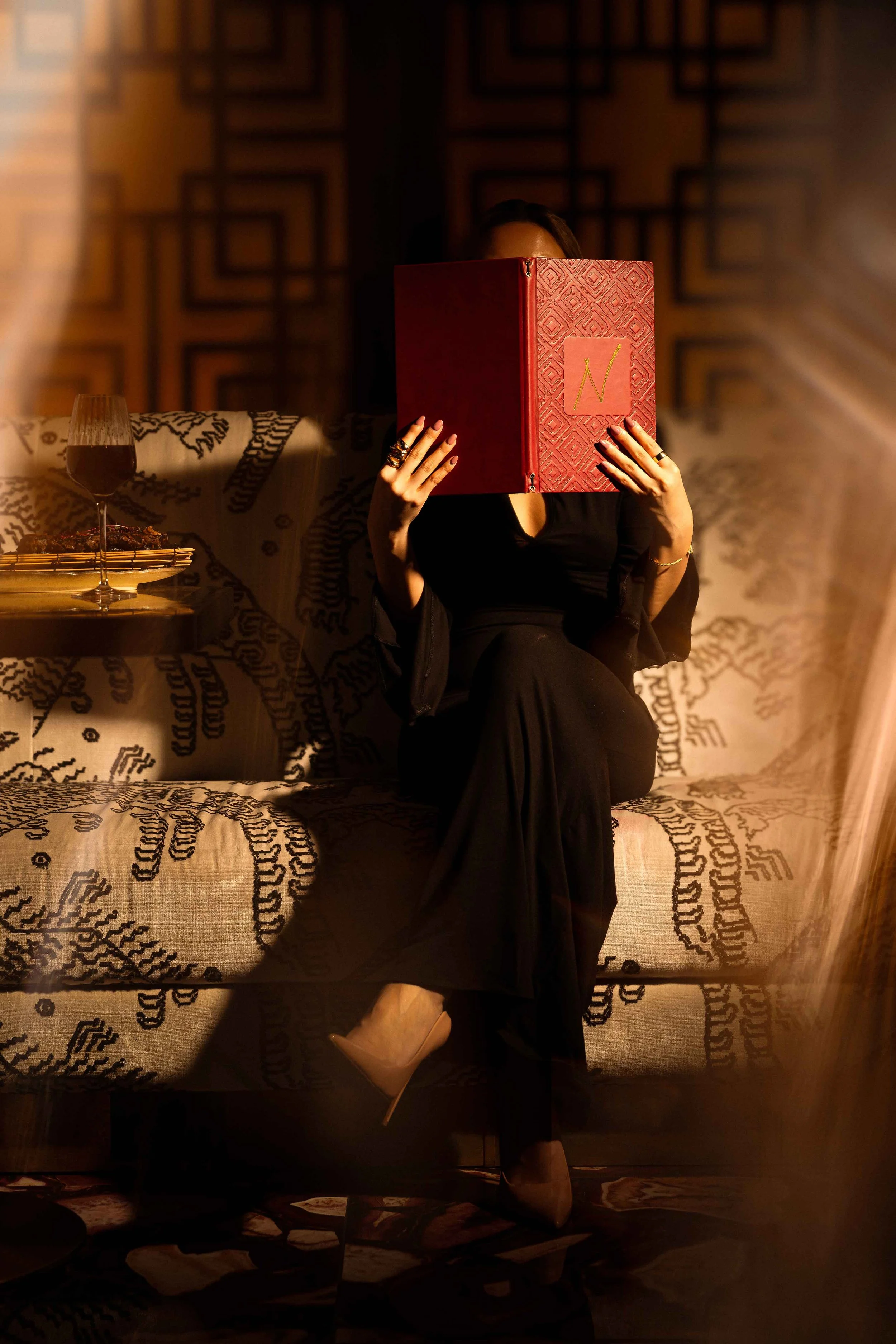 Person sitting on a sofa, holding a large red book in front of their face. They are wearing black clothing and beige high heels. A glass of red wine and some food on a plate are on a side table. The room has dim lighting and wooden geometric wall decor.