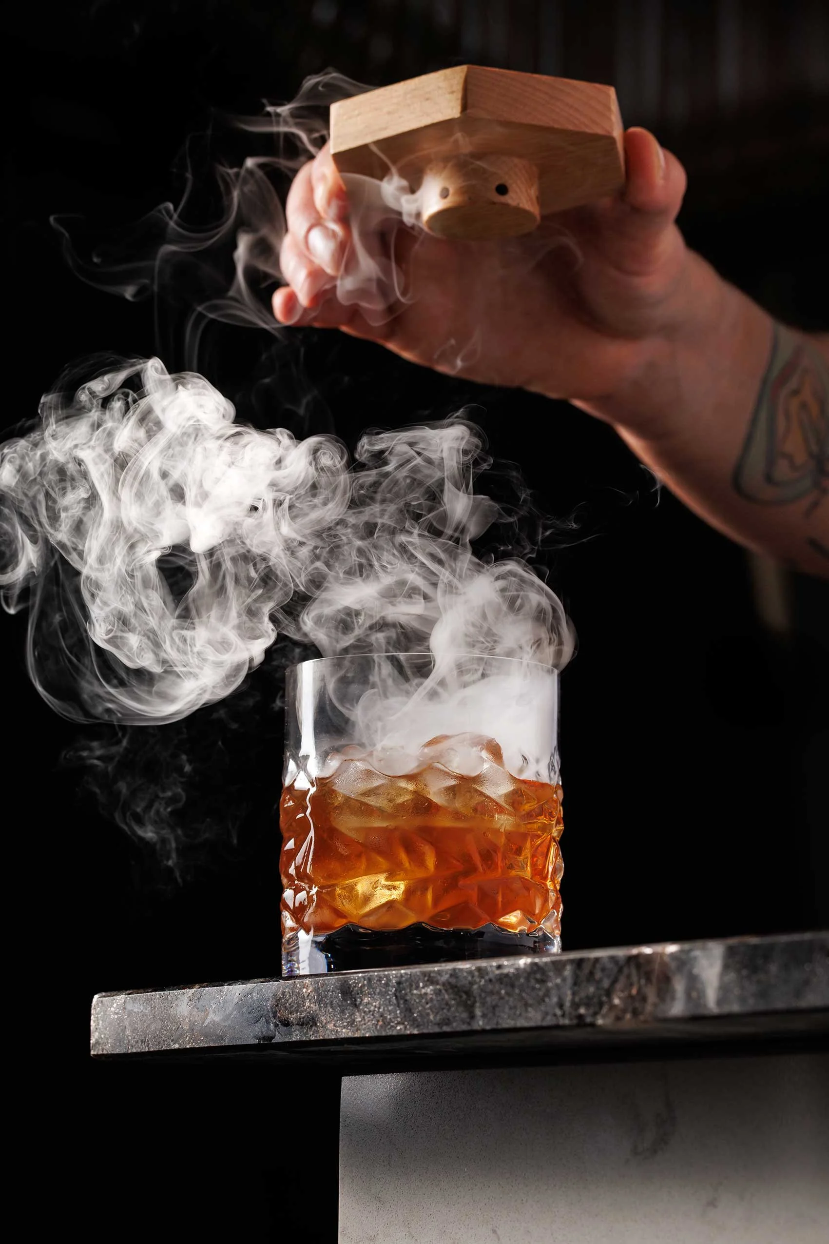 A hand holding a wooden device over a glass of amber-colored drink with ice, with smoke or vapor swirling around the glass against a dark background.