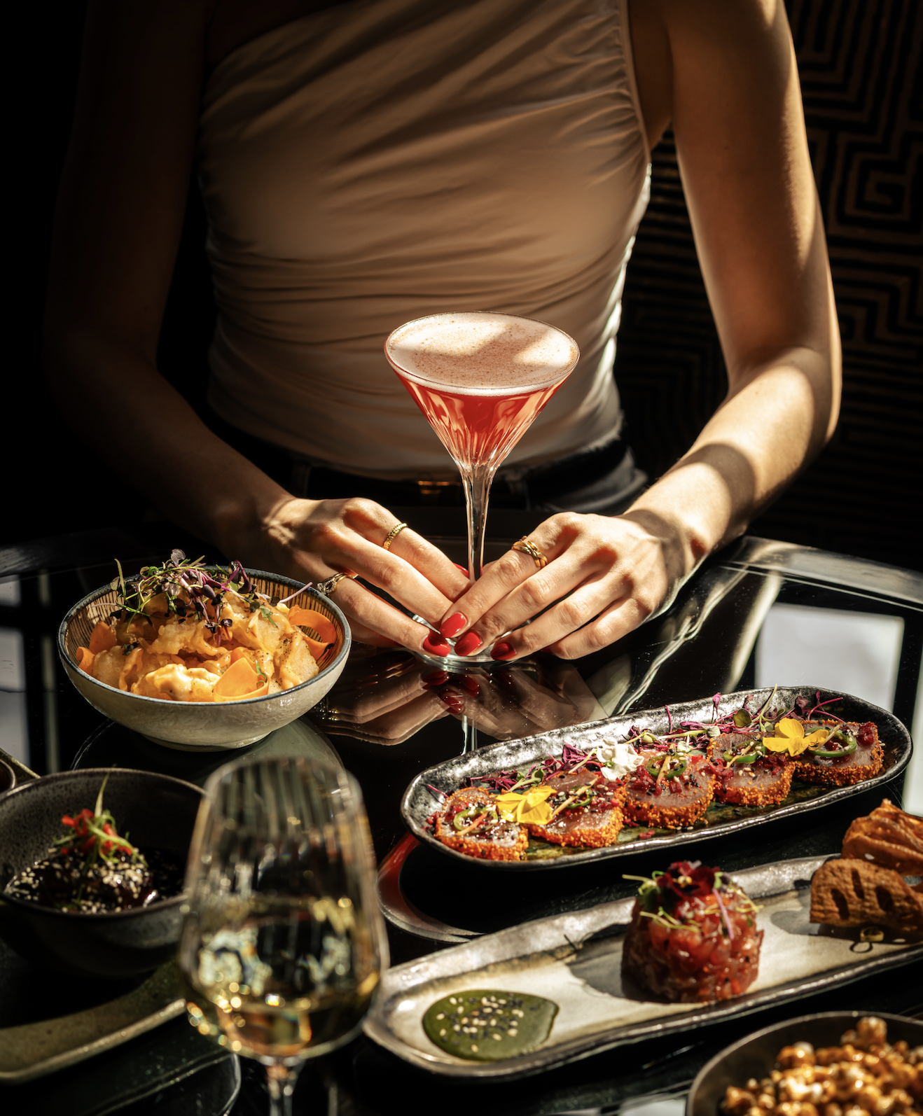A person with red nail polish and multiple rings on their fingers holds a pink cocktail glass filled with a foamy drink. On a table, there are plates of gourmet food including a bowl of pasta, a platter of sliced meat with garnishes, small appetizers, and a glass of white wine.