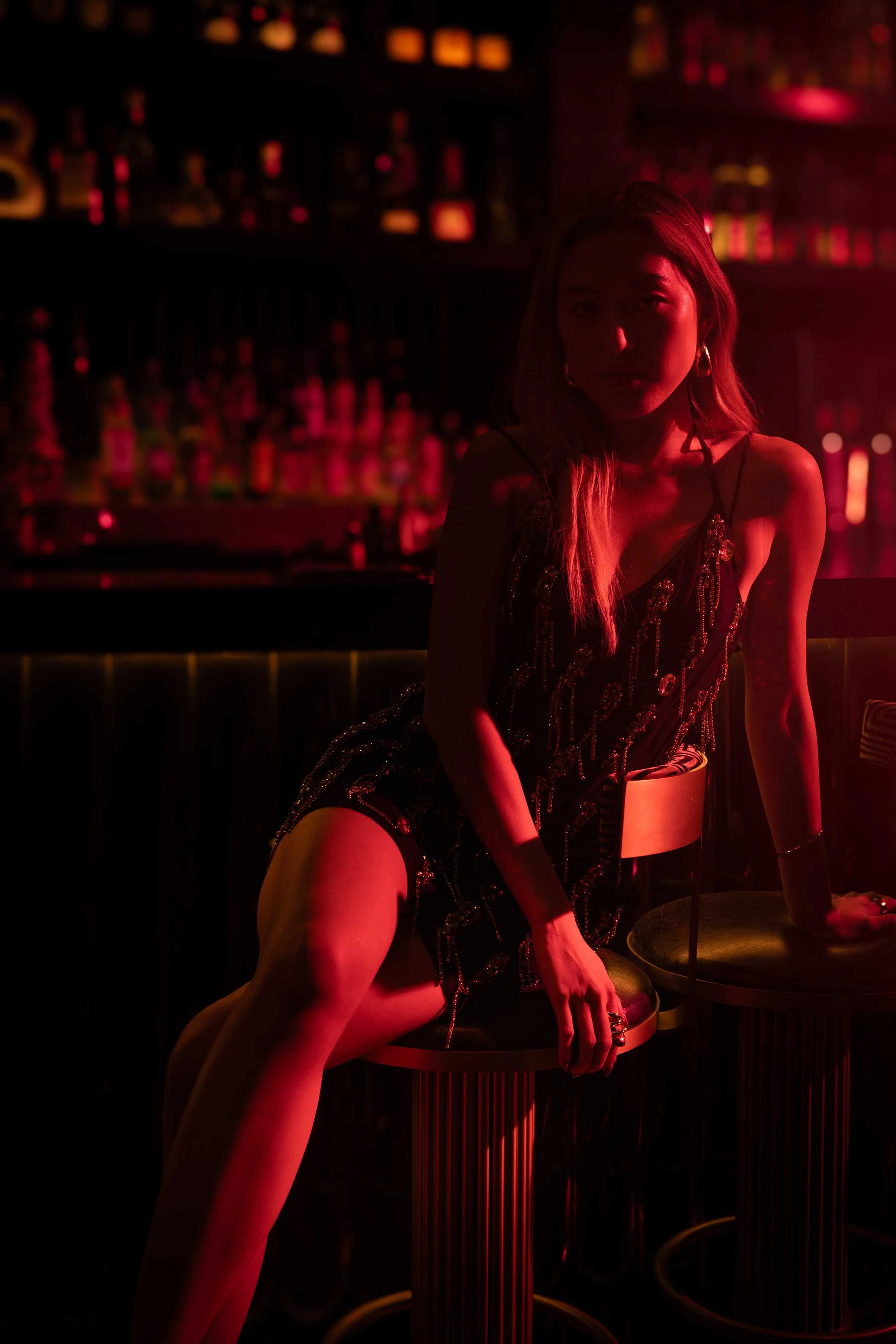 A woman in a black dress with sequins, sitting at a bar in a nightclub with red lighting and bottles visible in the background.
