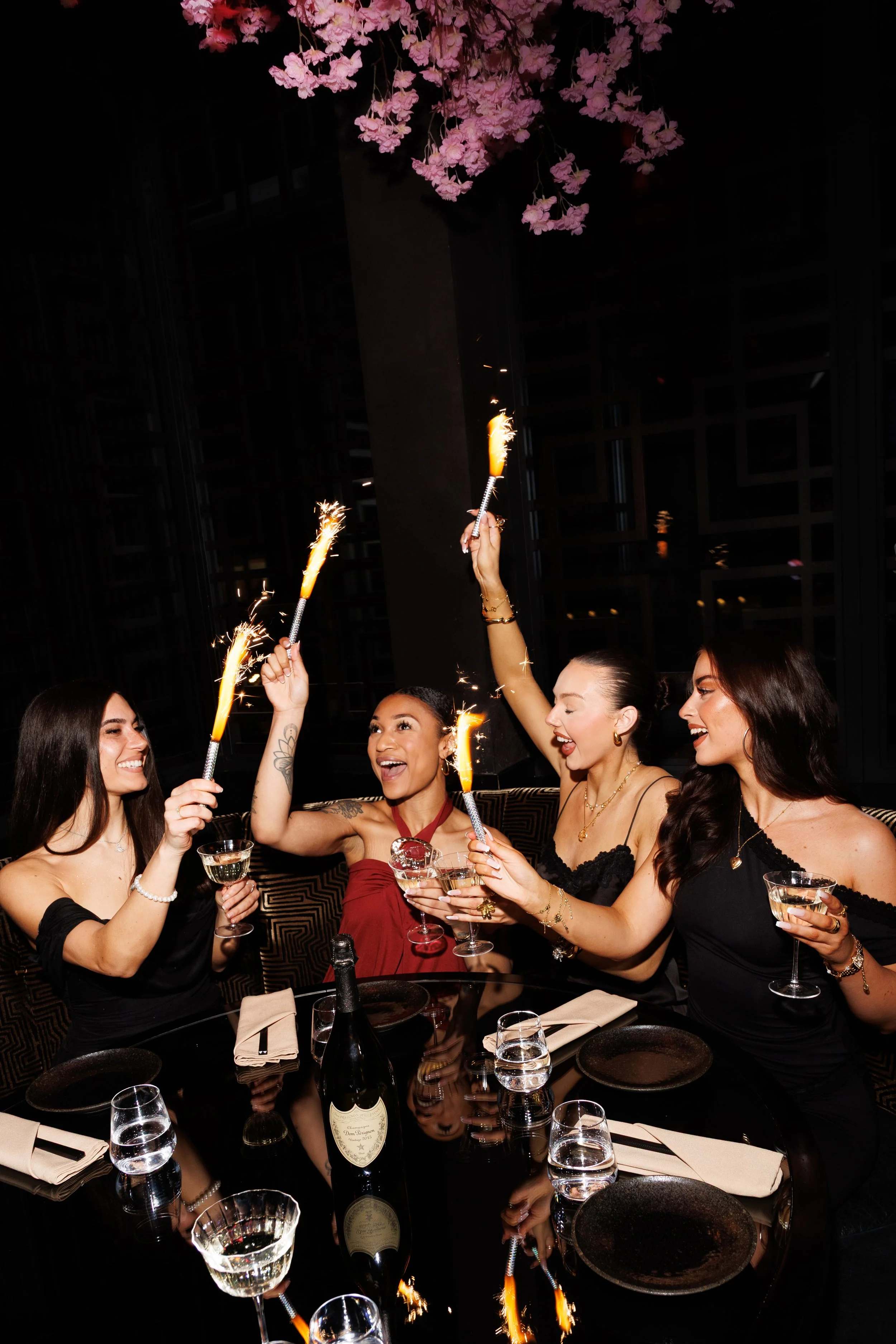 Five women celebrating at a party, holding sparklers and champagne glasses, with a birthday cake in the background and a dark, elegant setting.
