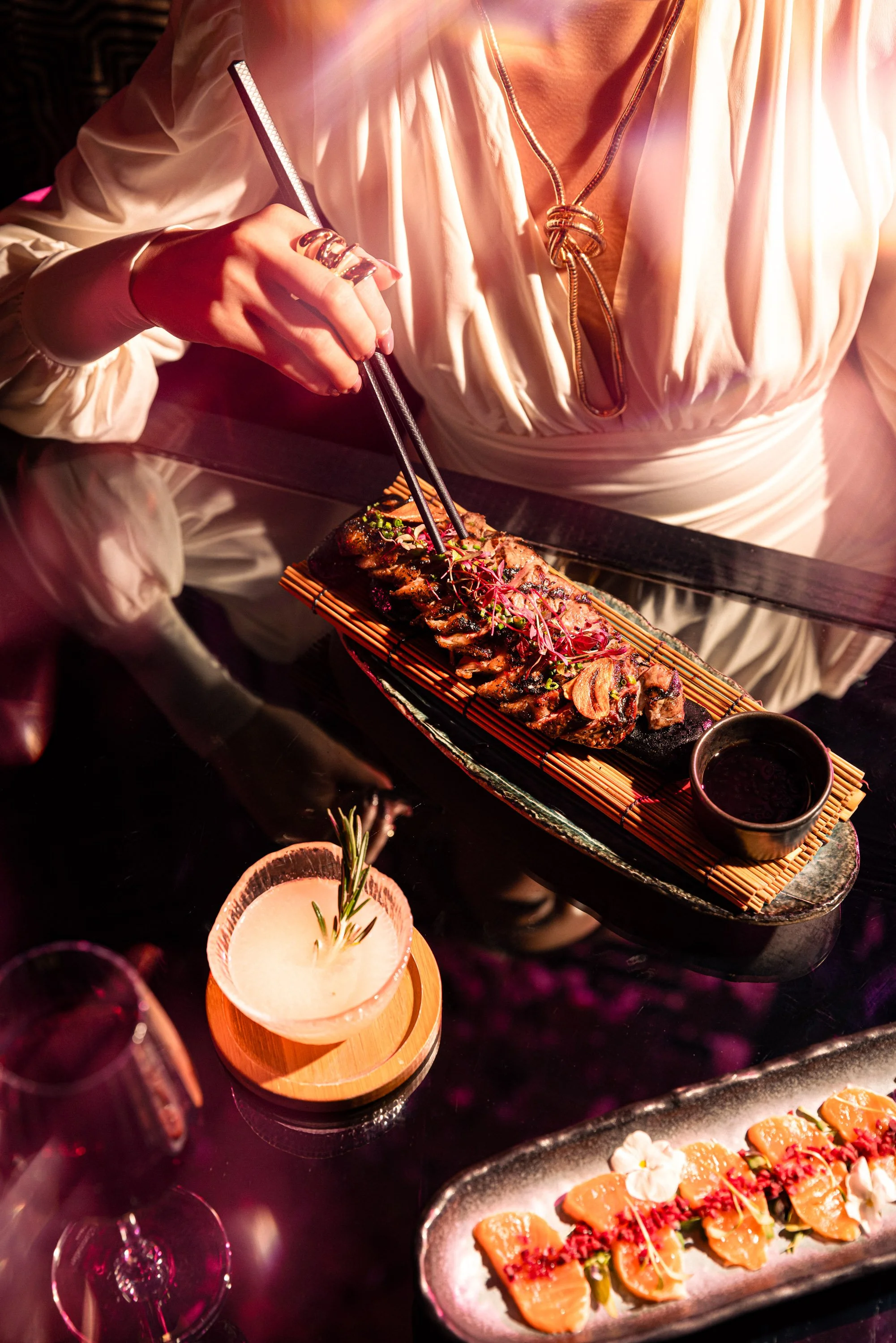 A person in a white dress with jewelry is using chopsticks to pick up a piece of sushi from a long black platter topped with garnishes. There are two cocktail glasses and a sliced lemon with herbs in the foreground.