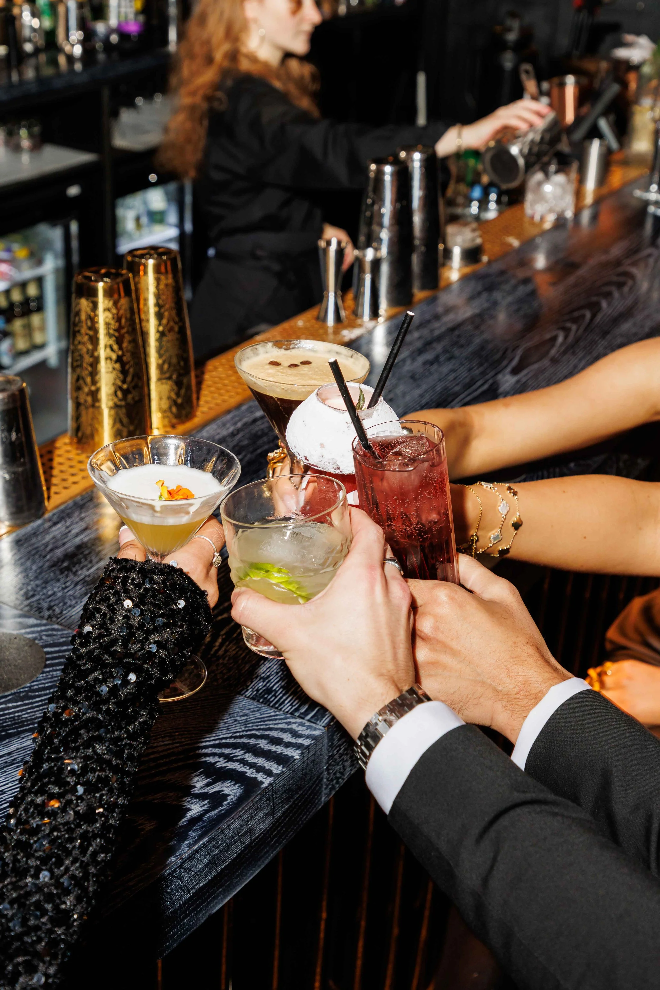 People raising glasses filled with cocktails for a toast at a bar with a variety of drinks in the background.