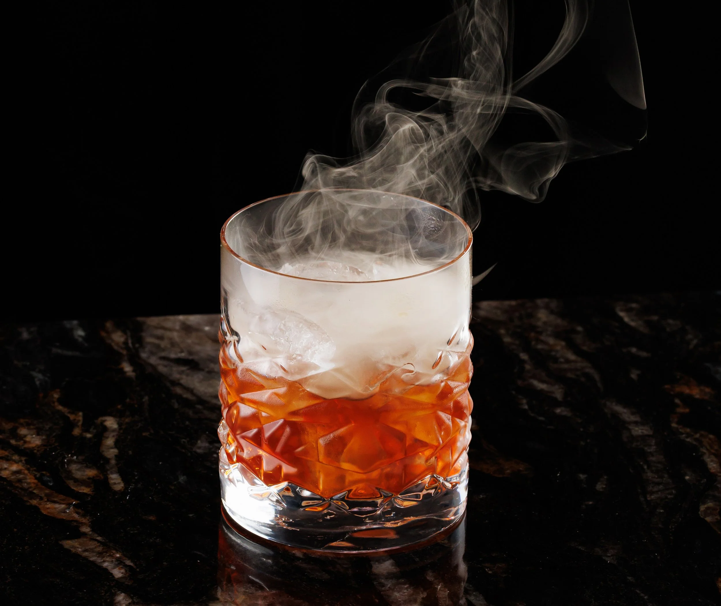 A glass with an orange hue pattern, filled with ice, on a dark marble surface, with smoke rising from it against a black background.