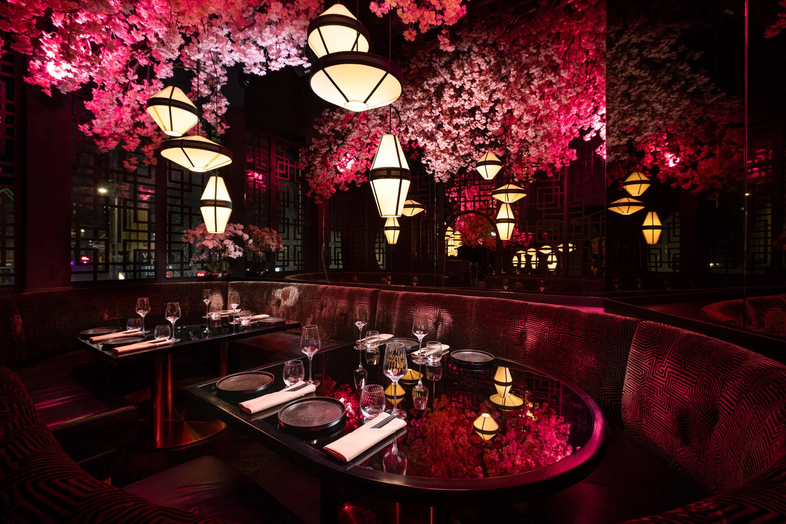 Elegant restaurant interior decorated with pink cherry blossom trees and hanging lantern-style lights, with a dining table set with glasses, plates, and napkins.