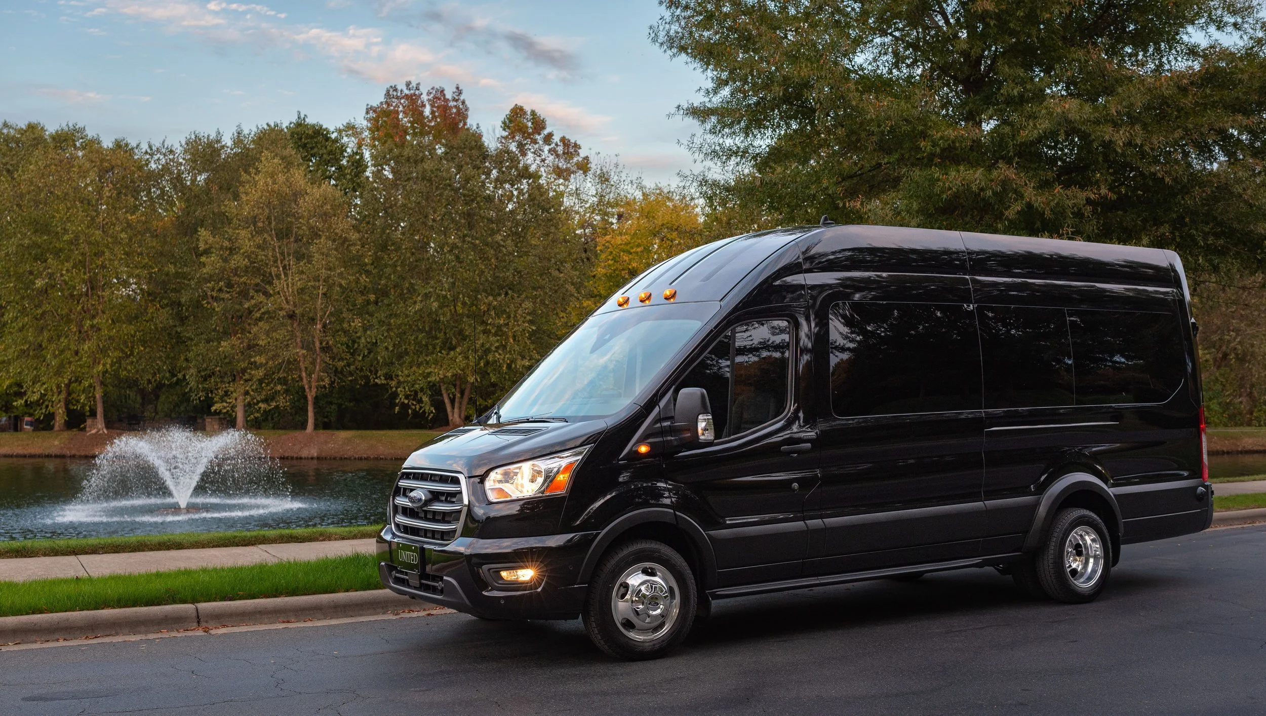 A black cargo van parked beside a pond with a fountain, trees in the background, and a partly cloudy sky.