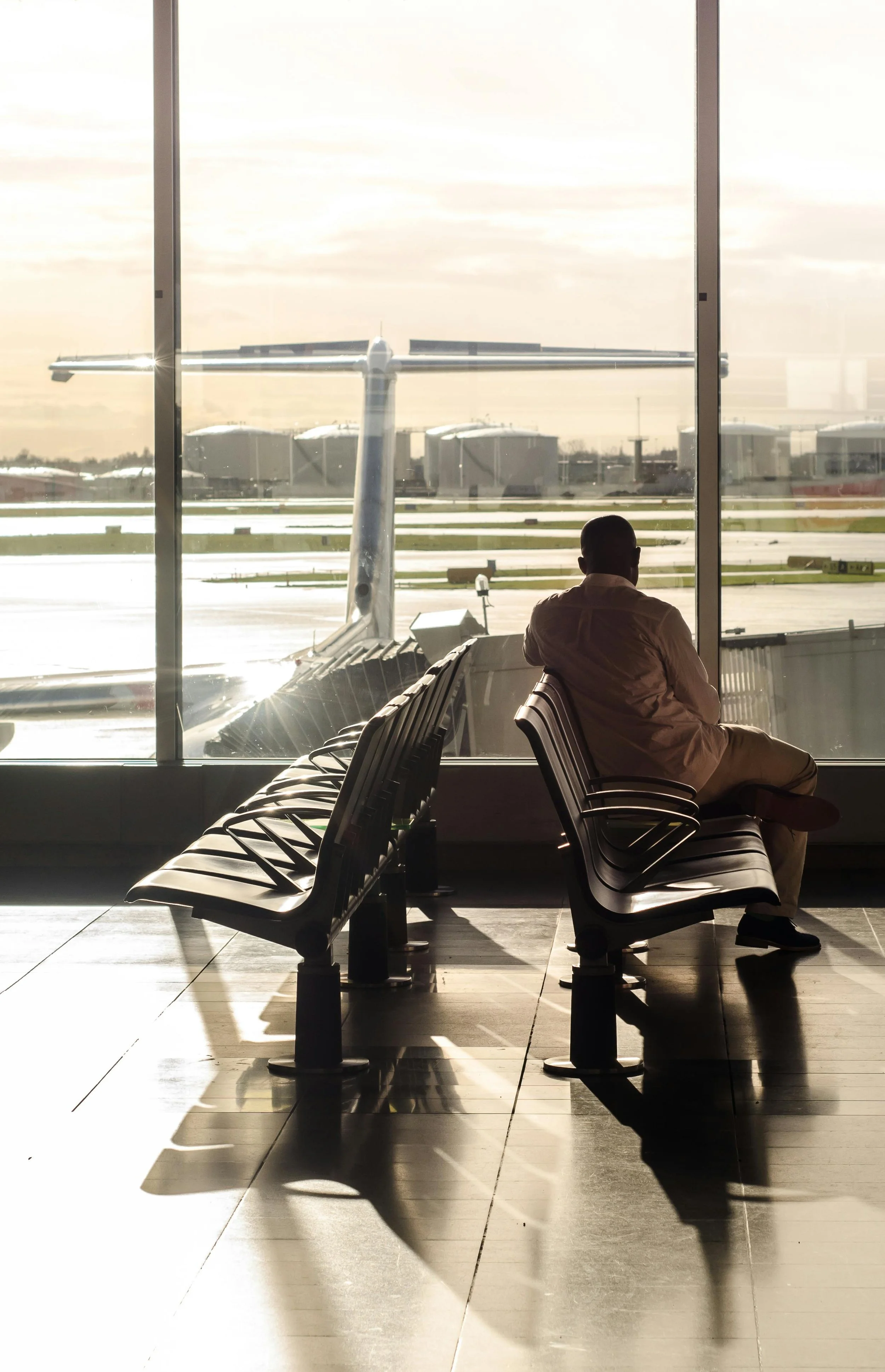 A man sitting alone in an airport waiting area, looking out the window at an airplane on the tarmac during sunset, with empty seats casting shadows.