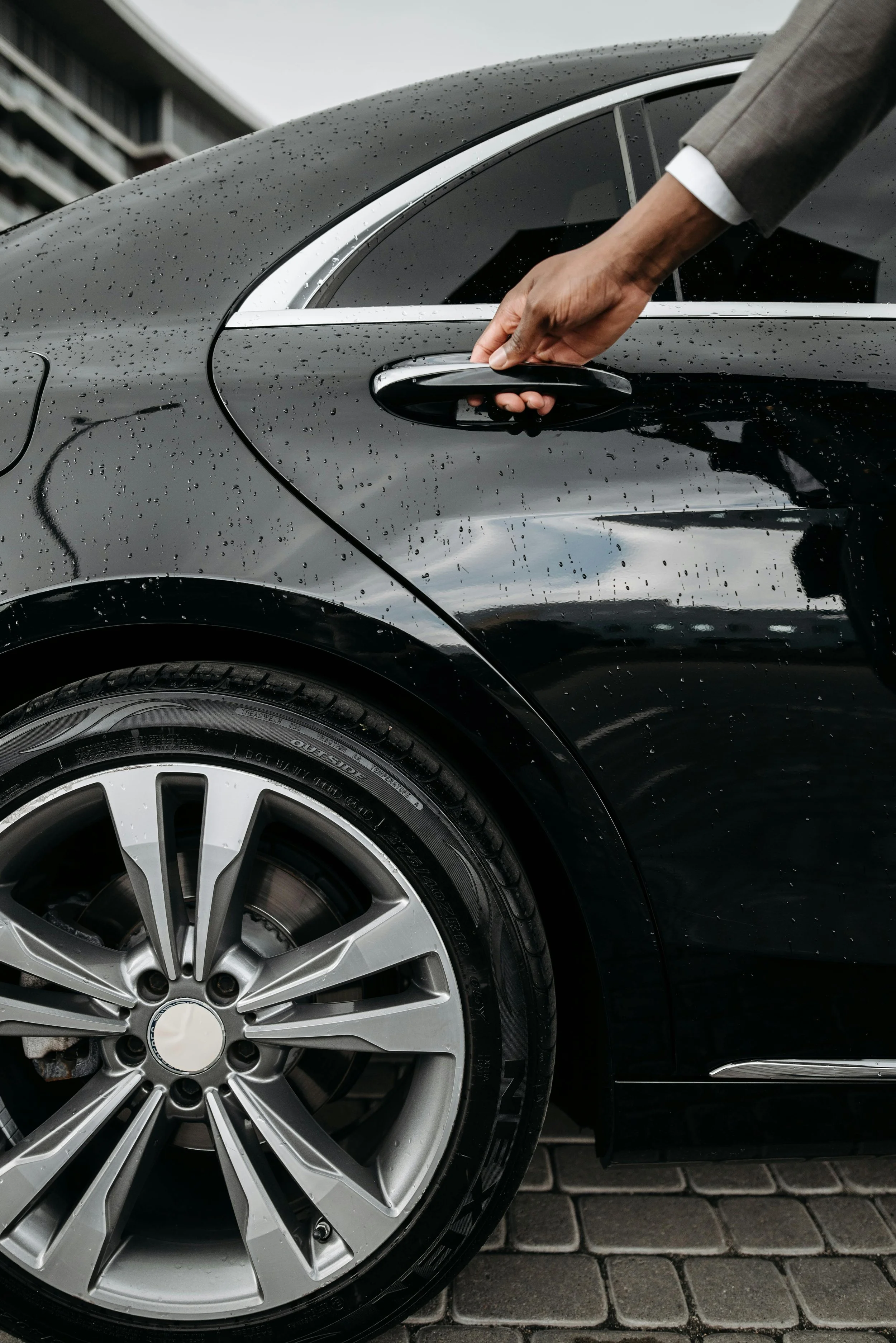 Close-up of black luxury car with water droplets on surface, showing part of the front wheel, silver rim, and car door handle being opened by a person wearing a grey suit.